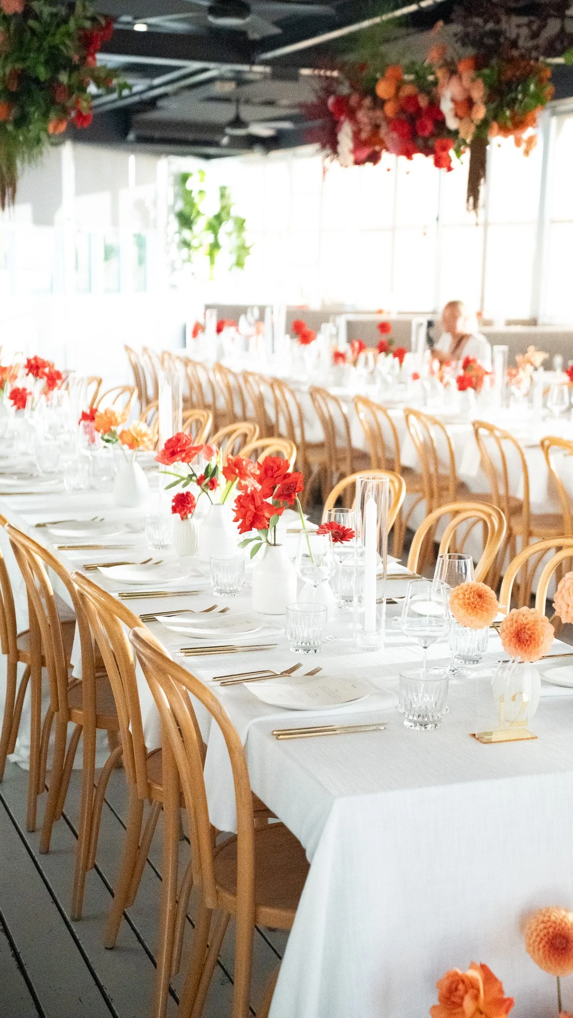 A banquet hall decorated with white tablecloths, floral centerpieces, and glassware, set up for a formal event with wooden chairs and hanging floral arrangements.