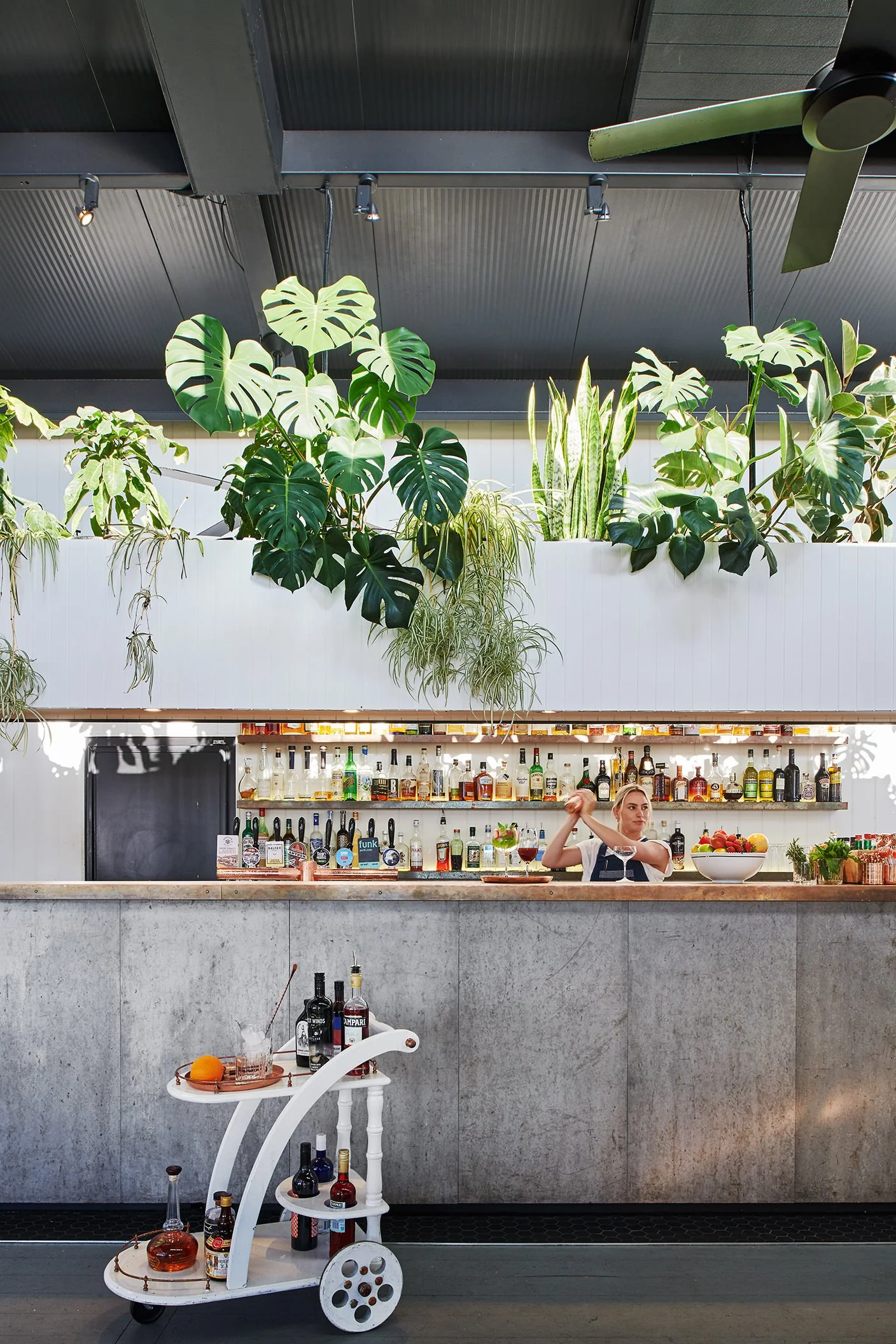 A bar area with a bartender mixing a drink behind a counter, with a white, multi-tiered cart in the foreground holding bottles of liquor, and tropical plants hanging above the bar in an indoor setting with modern decor.