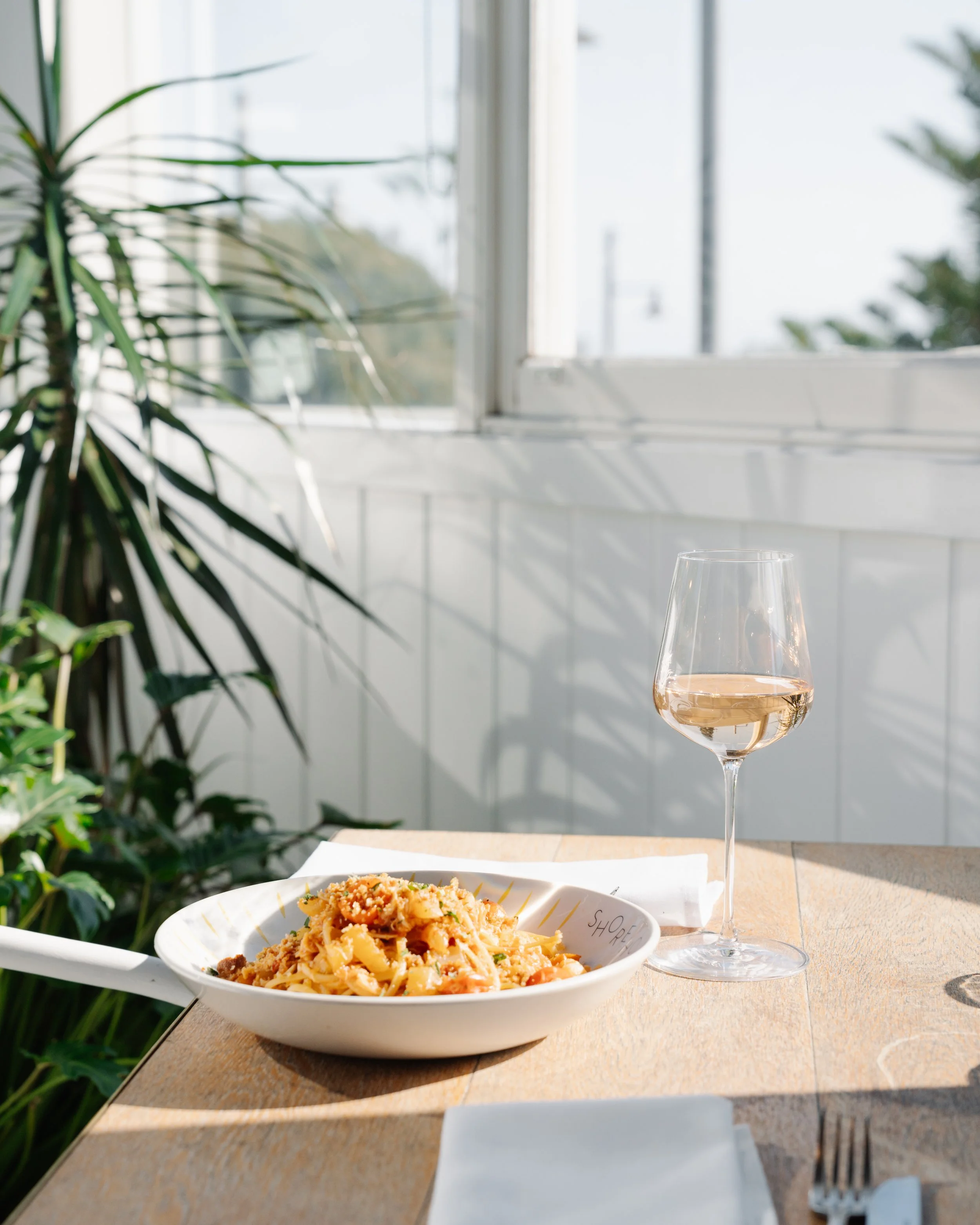 A bowl of pasta with a glass of white wine on a wooden table near a window with sunlight and greenery outside.