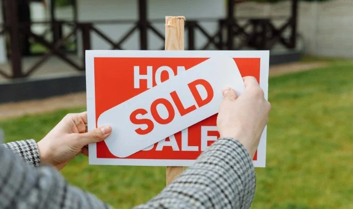 Person putting a 'SOLD' sign over a 'HOUSE SALE' sign outside on a lawn.