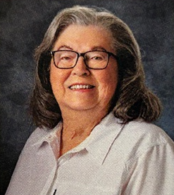 A portrait of an elderly woman with shoulder-length gray hair, glasses, wearing a white shirt, smiling at the camera.