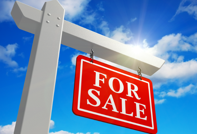 White signpost with a red "For Sale" sign hanging from it against a blue sky with clouds.
