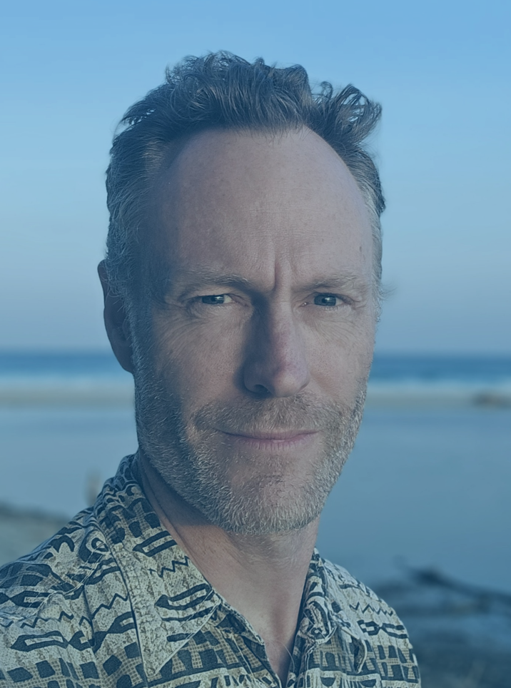 Profile photo of Jonathon Carle wearing a patterned shirt and smiling slightly with a beach, ocean and sky visible in the background.