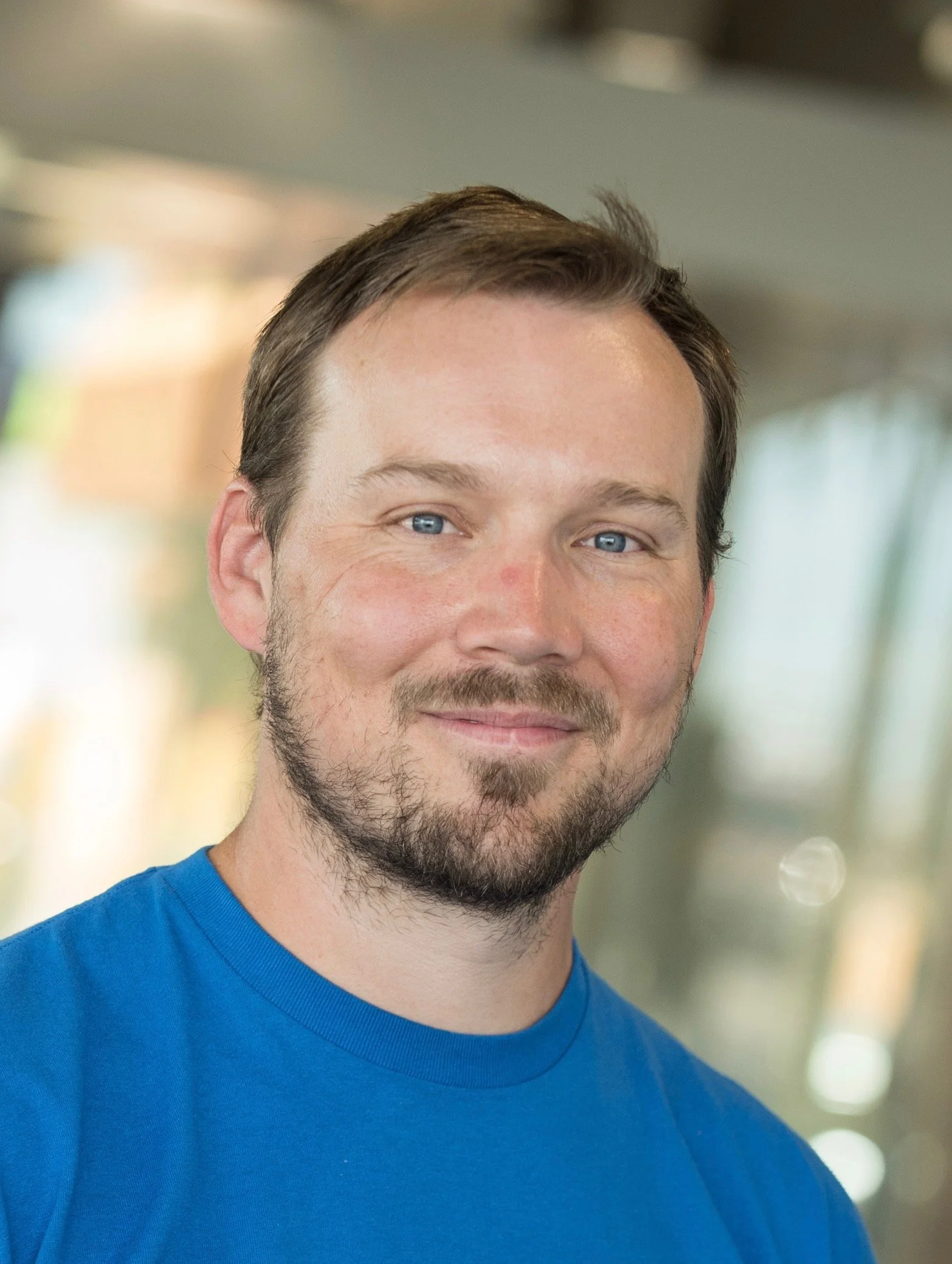 A man with short brown hair, blue eyes, and a beard, wearing a blue t-shirt, smiling at the camera in an indoor setting with blurred background.