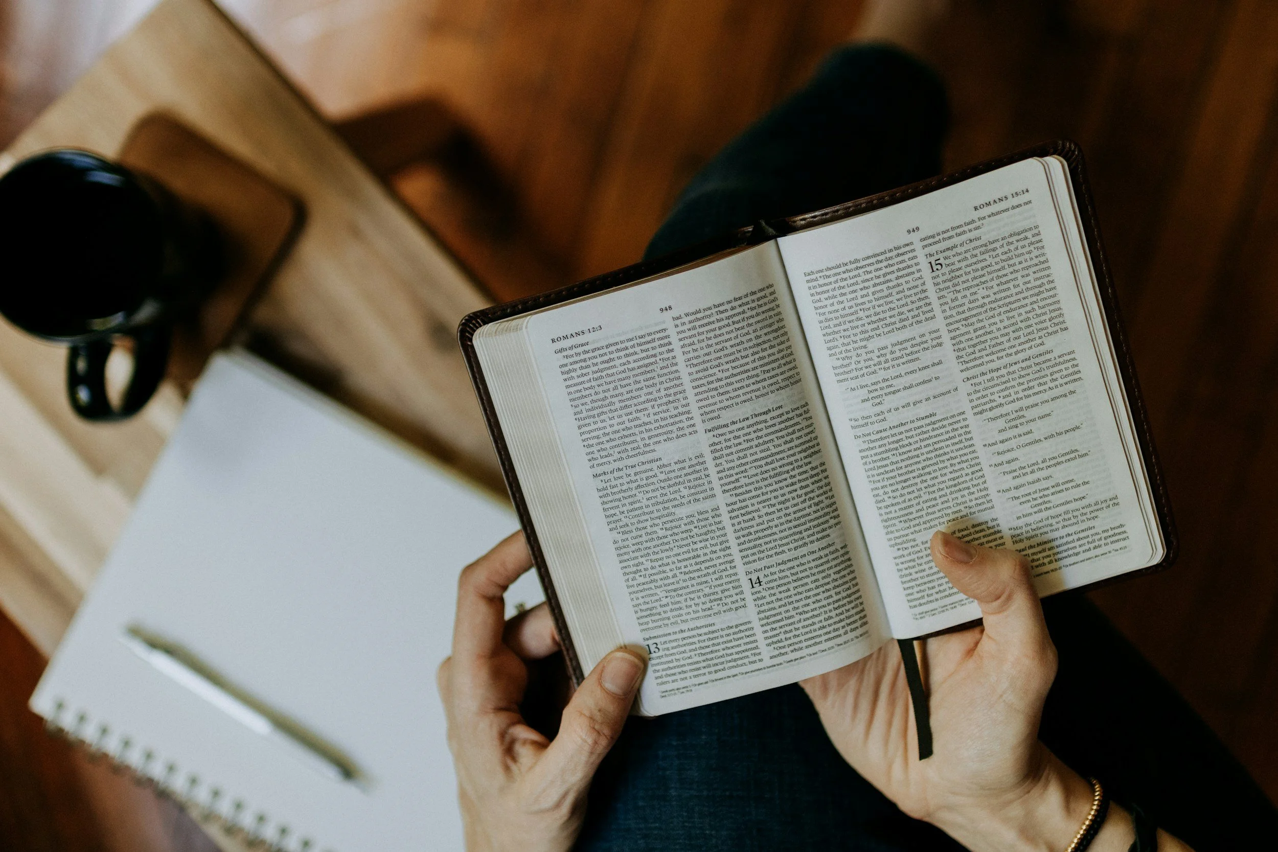 Person reading a Bible with a black coffee mug on a wooden table.