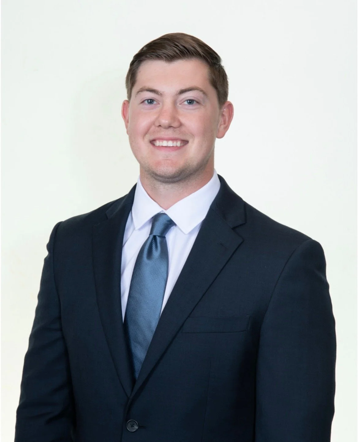 A young man in a black suit, white shirt, and blue tie, smiling at the camera with a plain light background.