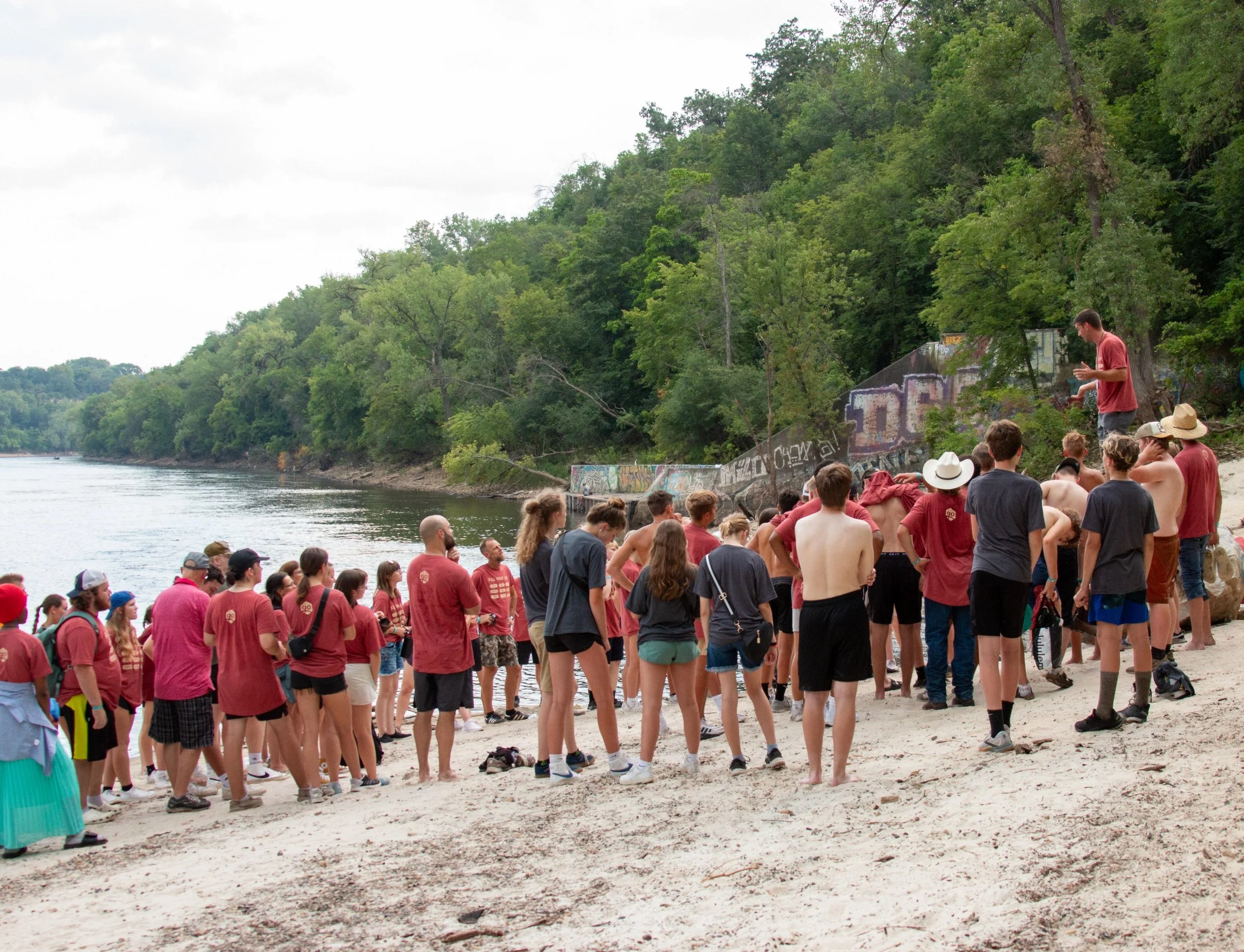 A large group of people gathered on a sandy beach near a river, with dense green trees on the opposite bank. Some individuals are wearing hats and casual clothing, and a man is standing on rocks above the crowd, speaking to them.