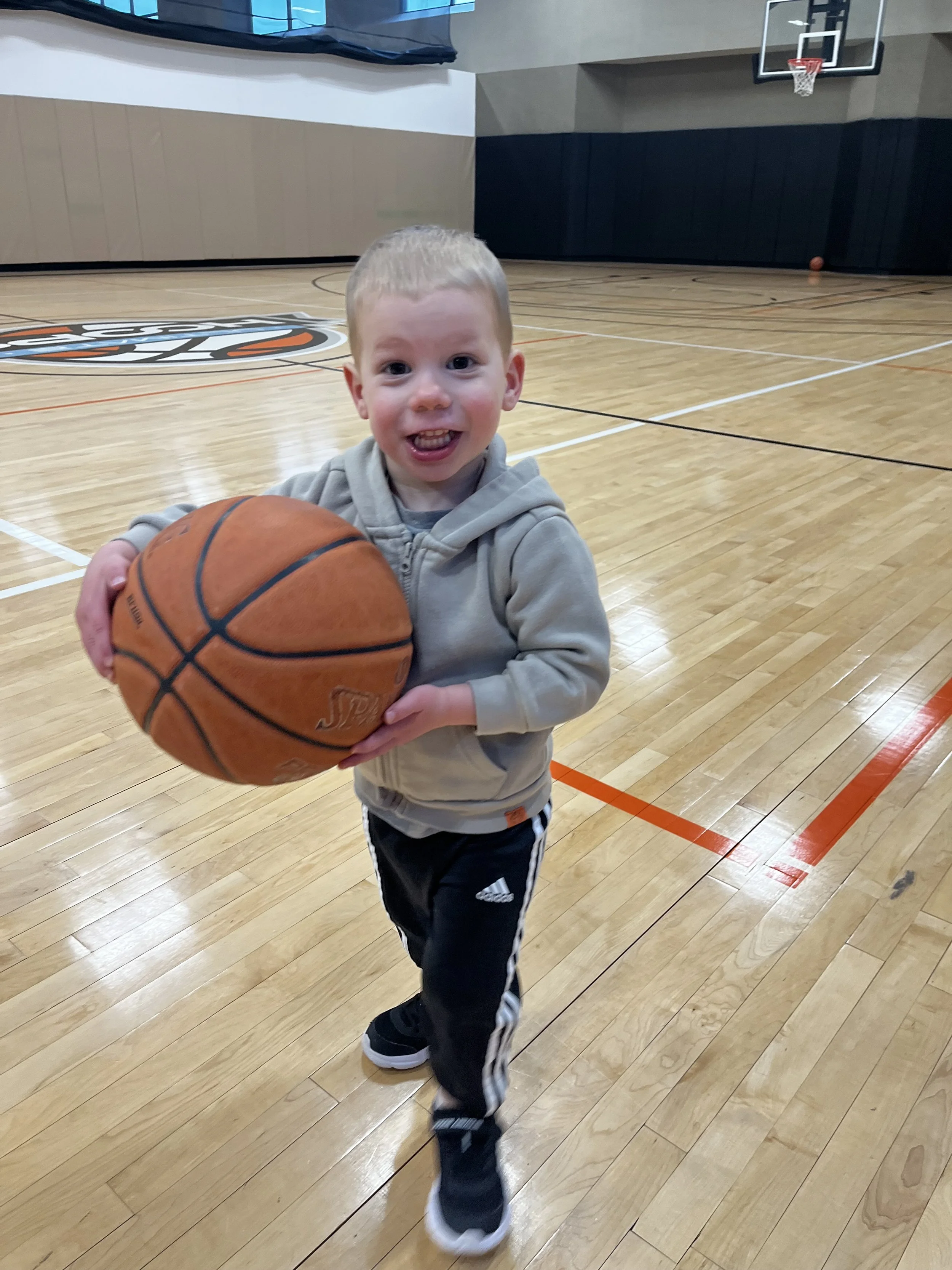 A young boy holding a basketball in a gymnasium, smiling and running towards the camera.