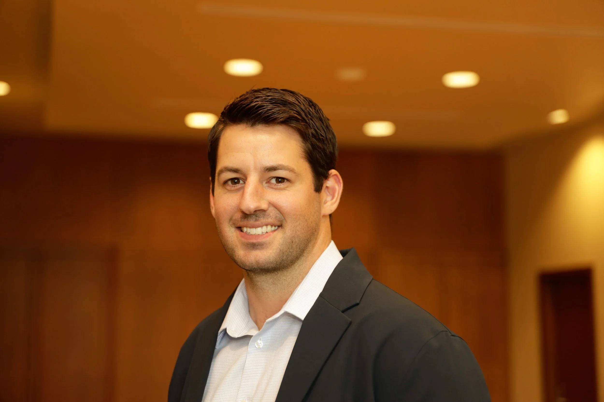 A man with dark hair wearing a suit smiling in an indoor setting with warm lighting.