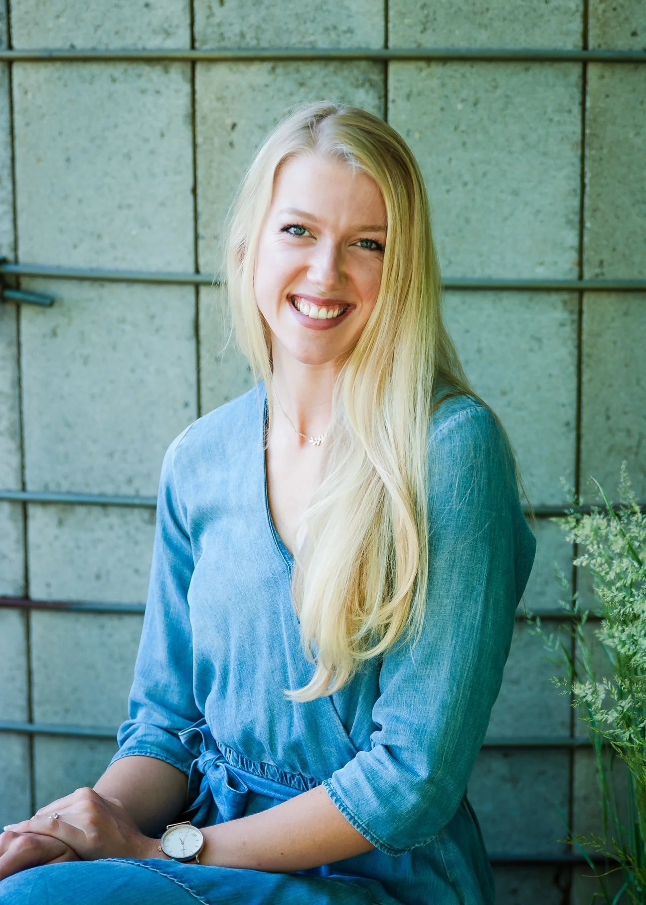 A smiling blonde woman wearing a denim dress sitting outdoors against a concrete wall with greenery nearby.