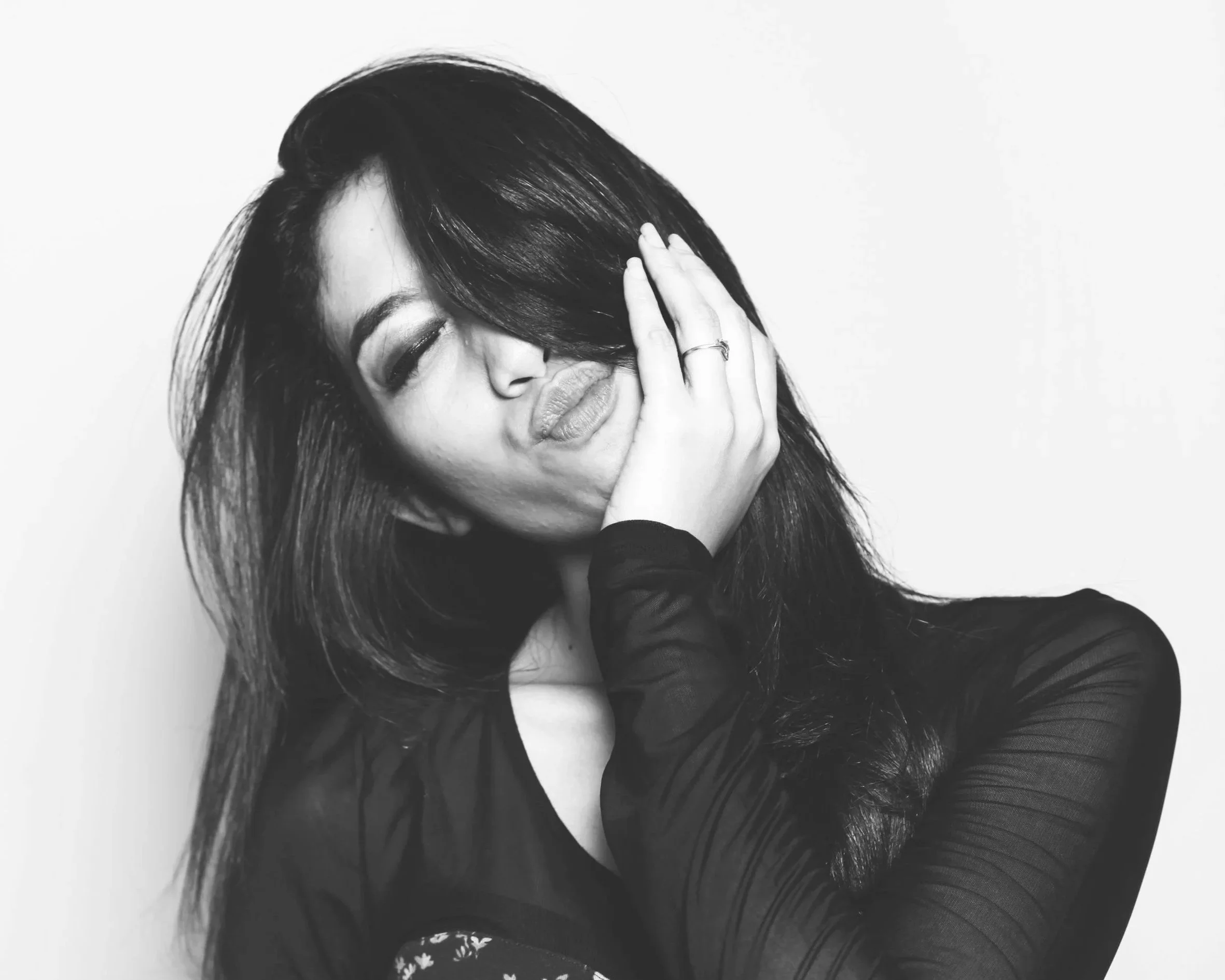 A black and white photo of a young woman with long dark hair covering part of her face, eyes closed, one hand resting on her face, lips slightly parted, wearing a dark long-sleeved top.