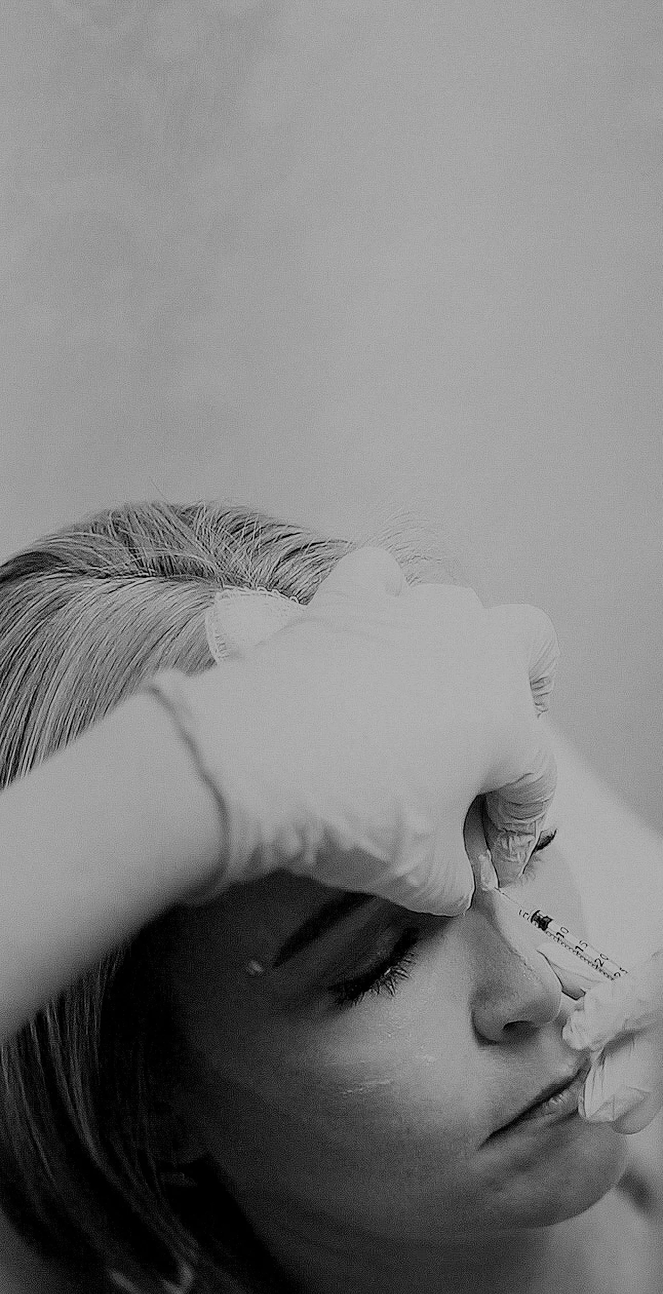 Black and white photo of a person receiving a cosmetic treatment, likely Botox or a similar injection, with a healthcare professional wearing gloves administering the injection to the person's forehead.