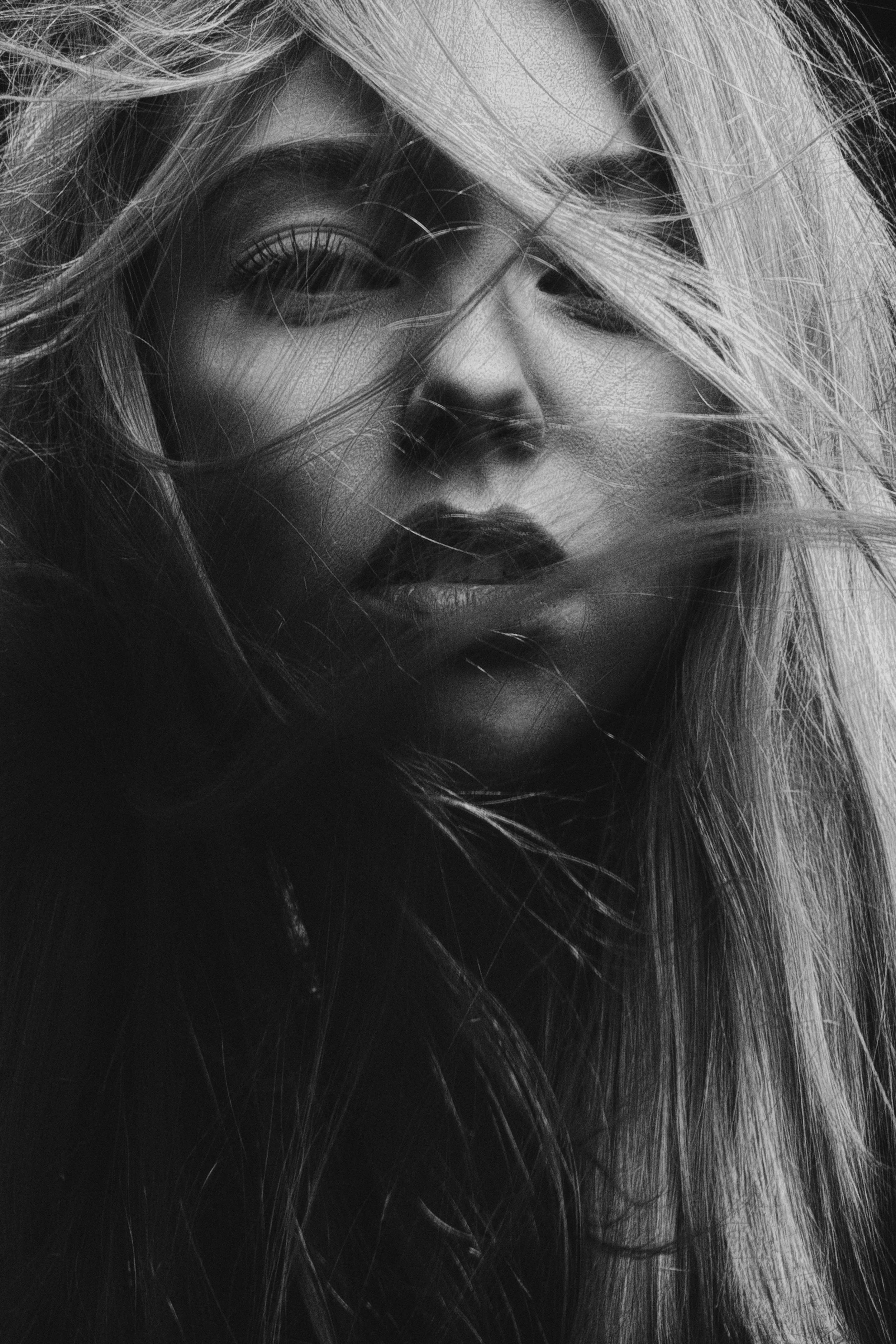 Close-up black and white photograph of a woman's face partially obscured by wind-blown hair, with intense expression and prominent eye makeup.