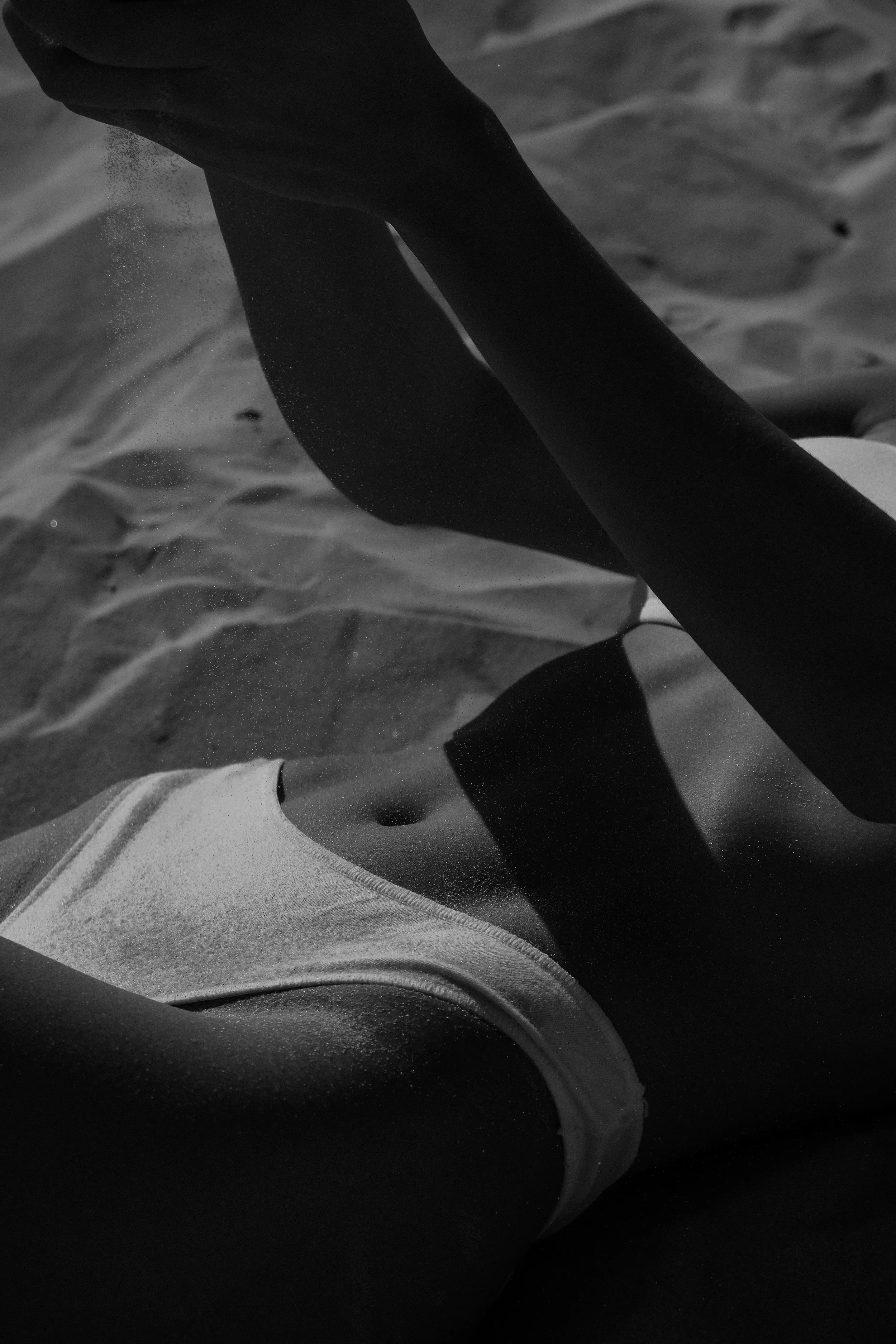 A black and white photo of a person's legs and feet in sandals on sandy ground, with shadows and textured sand visible.