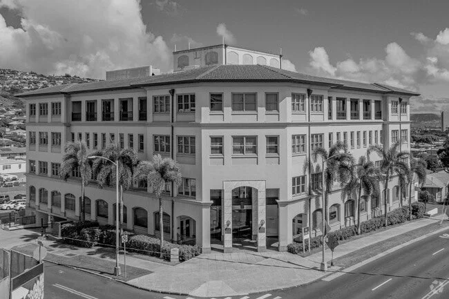 A large multi-story building with arched windows and palm trees lining the sidewalk in front. The building has a central entrance and a rooftop structure.