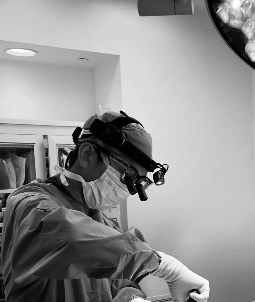 Dr. Justin Cheng wearing surgical scrubs, a mask, and magnifying loupes while performing plastic surgery in an operating room.