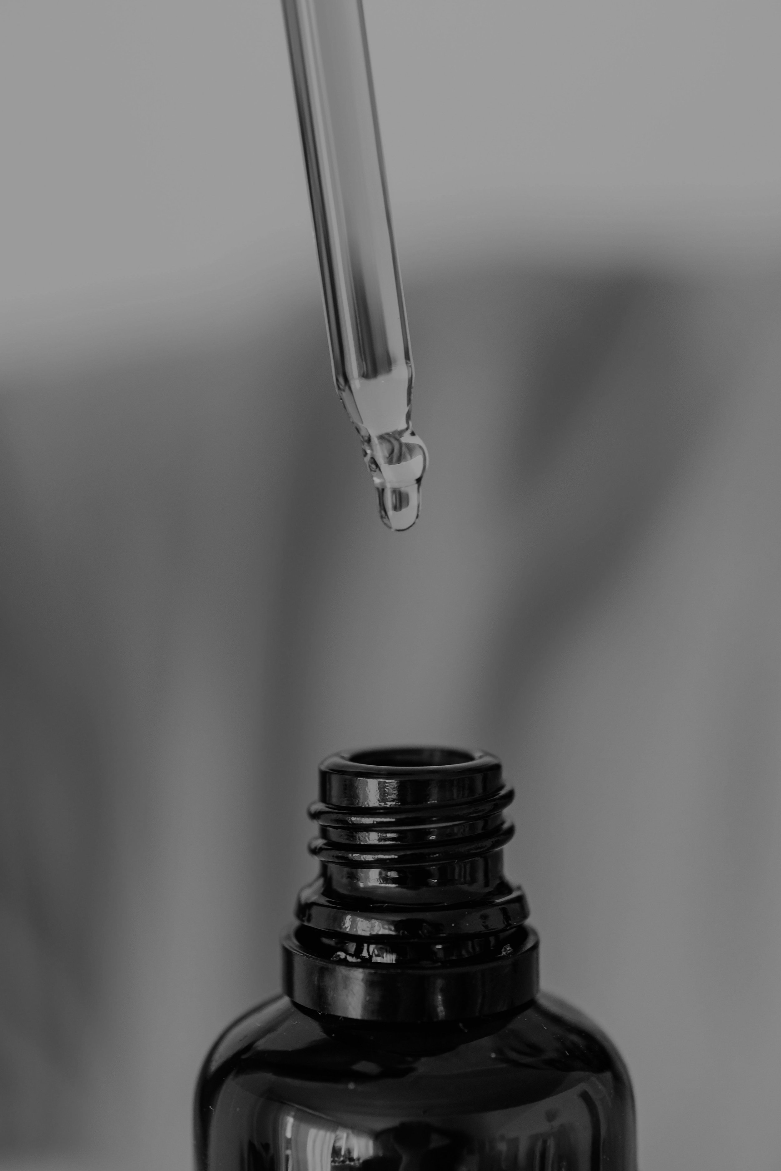 A close-up black and white photograph of a drop of liquid falling from a thin tube into a small container, with the focus on the droplet and tube.