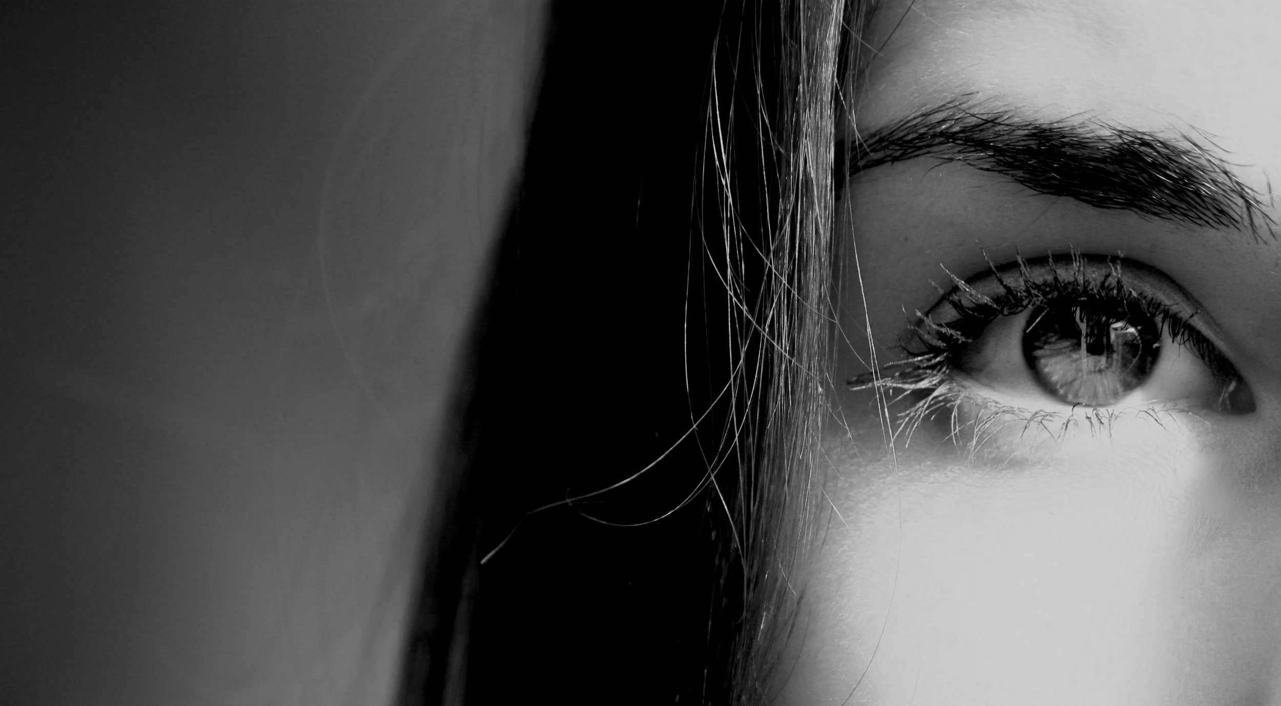 Close-up of a person's eye and eyebrow, with some hair falling over the person's face, in black and white.