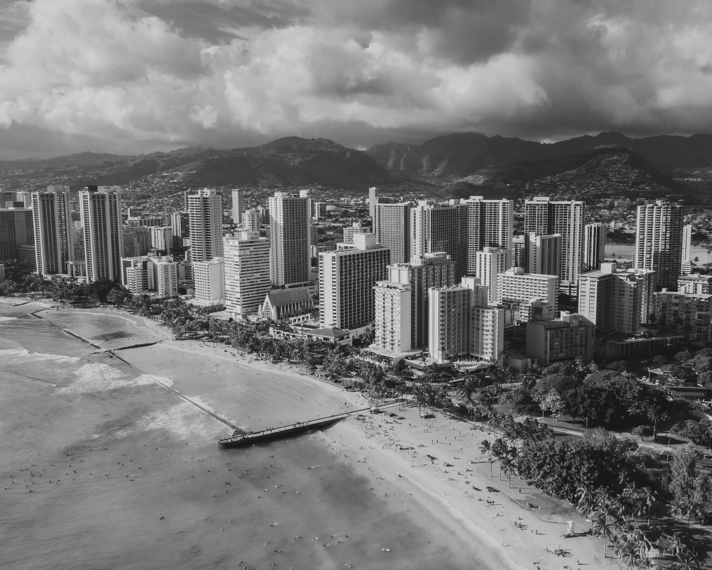 A black and white aerial view of a city with tall buildings, mountains in the background, and a coastline or beach area in the foreground.