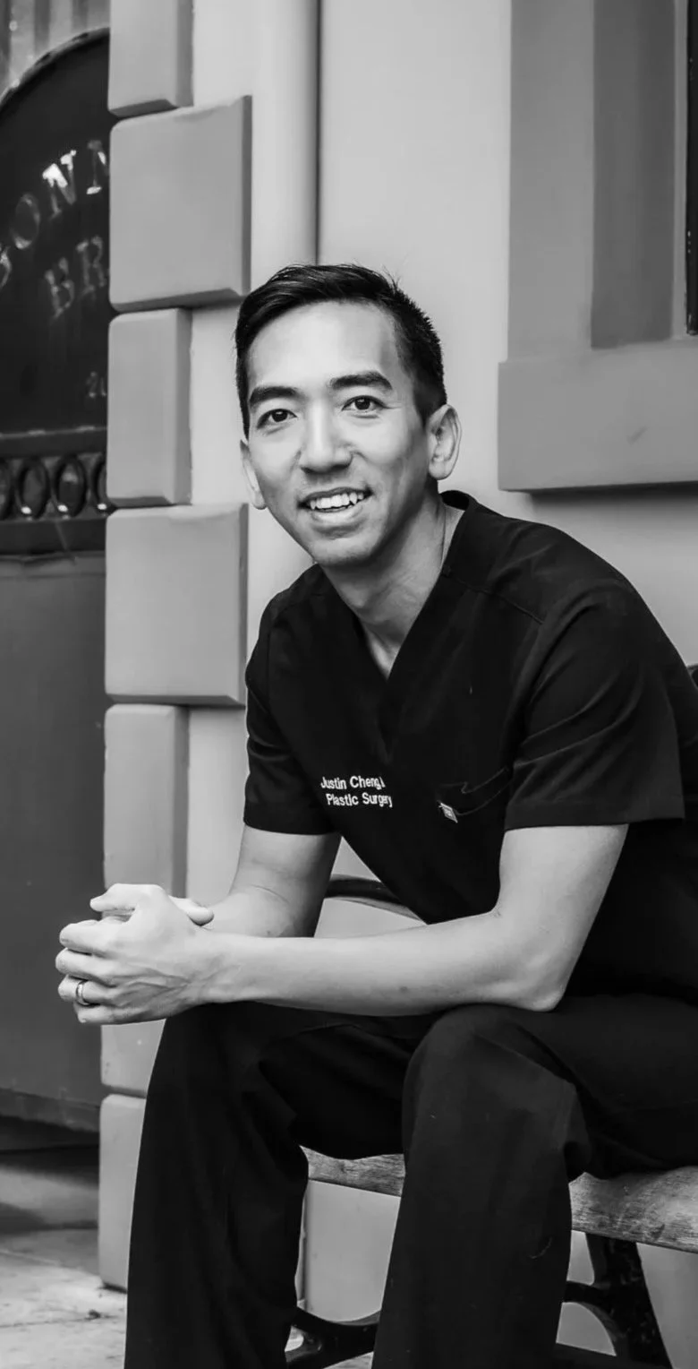Dr. Justin Cheng, Honolulu plastic surgeon, smiling in black scrubs in a modern clinic setting.
