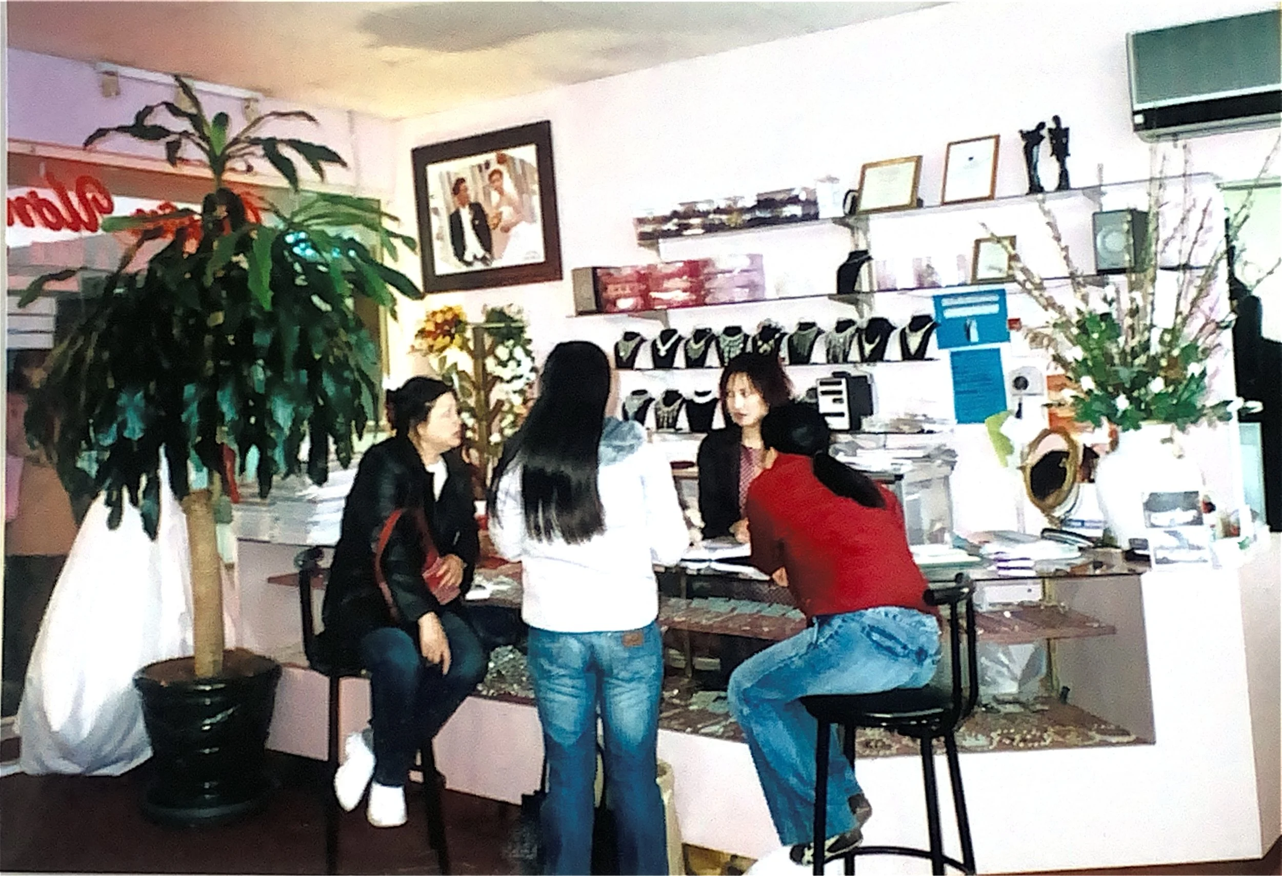 Four women gathered around a cluttered table in a cozy, decorated room with plants, artwork, and shelves with jewelry or small items, engaging in conversation.