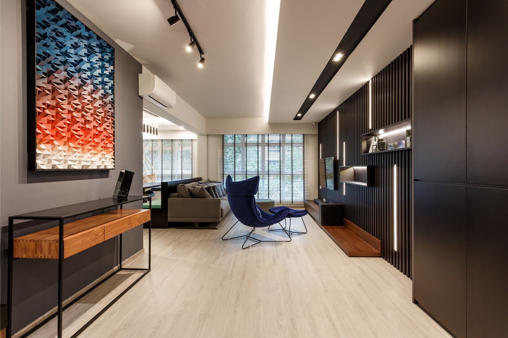Modern living room with gray and black furniture, a large wall-mounted TV, a blue lounge chair with a matching ottoman, a gray sofa, decorative wall art, wood and black accents, and large windows with blinds.