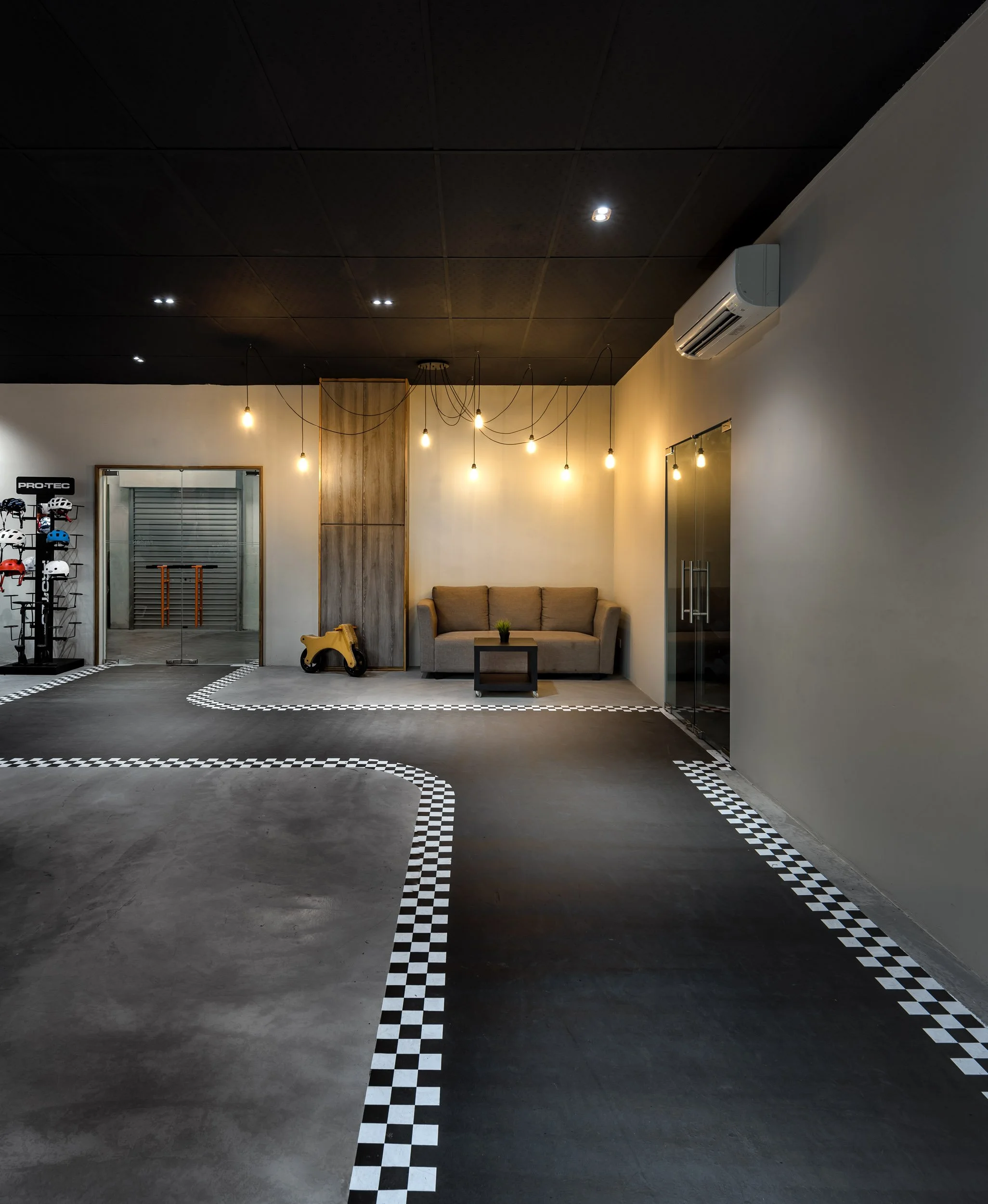 Interior of a modern room with black ceiling, beige walls, hanging light bulbs, a beige sofa, a small coffee table, and a corner with a toy ride-on car. There is a glass door, an air conditioner, and a display with helmets in the background.