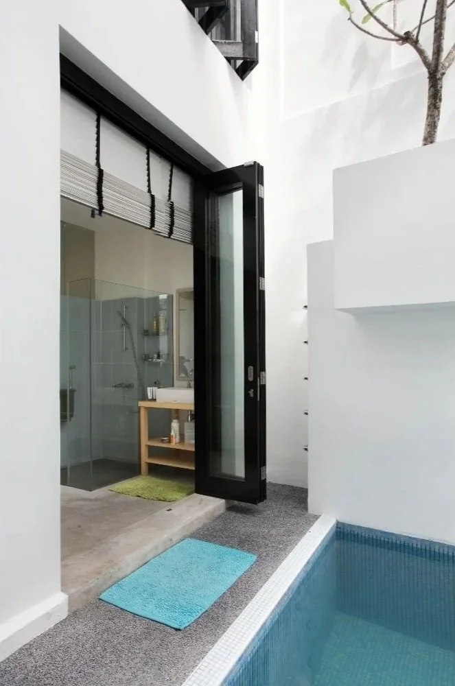 Open glass door leading from pool area into modern bathroom with walk-in shower and wooden vanity.