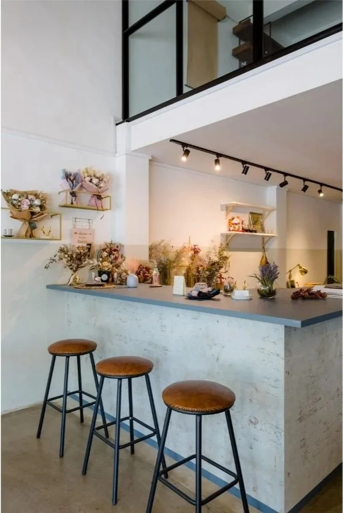 A minimalist indoor cafe with a white textured counter, three brown leather stools, and decorative items like dried flowers and small shelves with plants and ornaments. Track lighting and an open loft area are visible.
