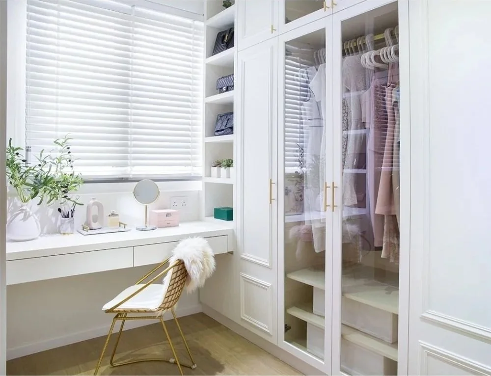 Bright, white bedroom with built-in wardrobe and window blinds. Small desk with mirror, plant, and decorative items. Gold chair with white cushion and fur throw.