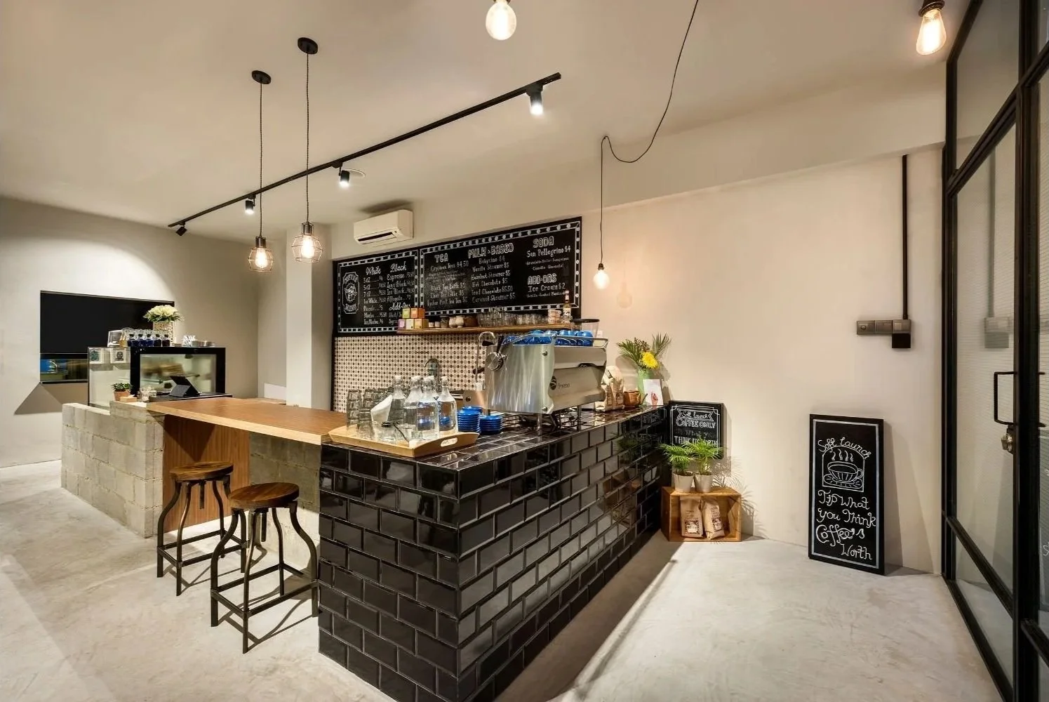 Modern coffee shop counter with black tiled front, wooden countertop, and stools. Menu board above, coffee equipment, and decorative plants; white walls and hung lightbulbs.