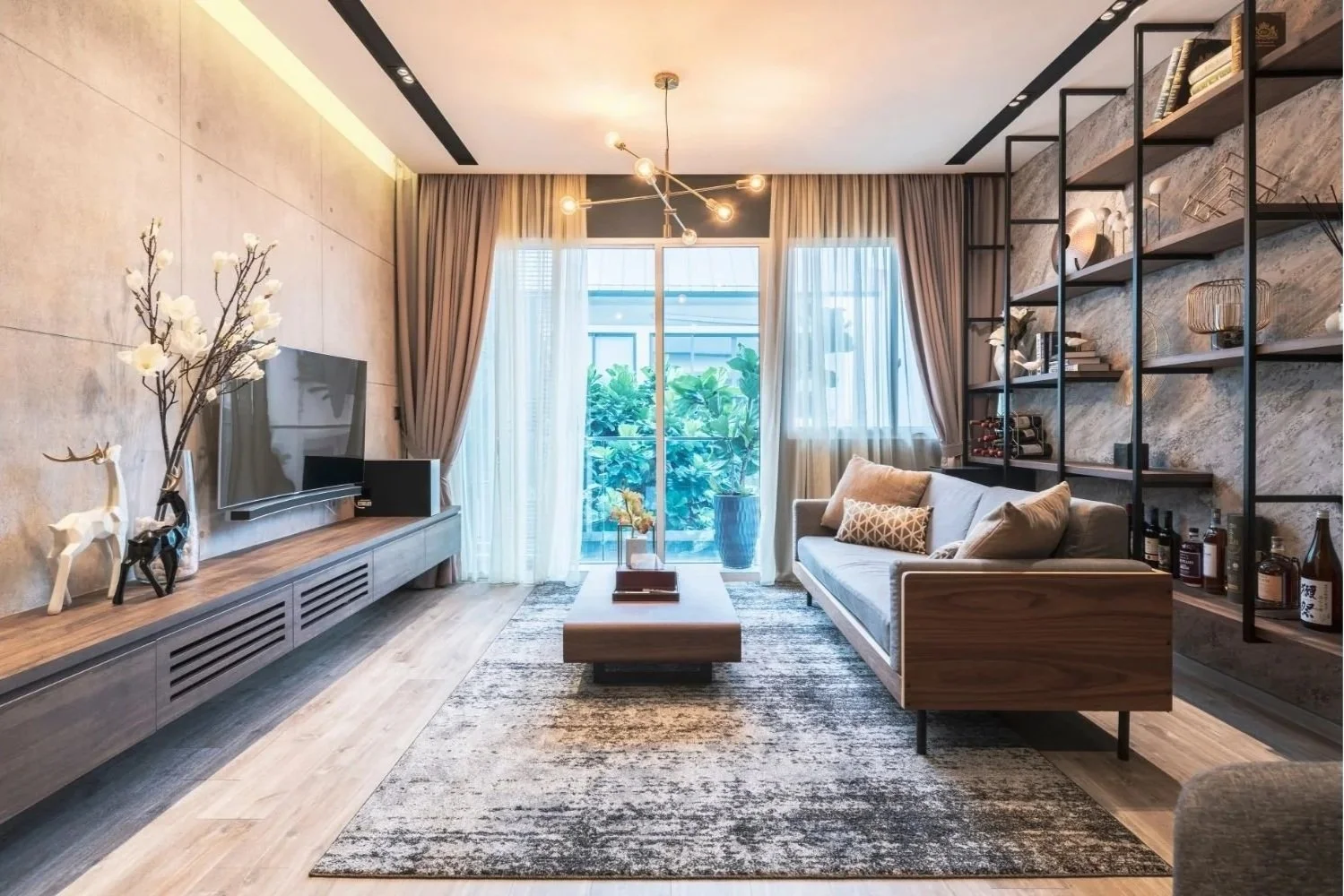 Modern living room with beige walls, wooden flooring, a gray sofa with pillows, a wooden coffee table, a TV on a long wooden console, a bookshelf with decor items, and large windows with curtains leading to a balcony with greenery.