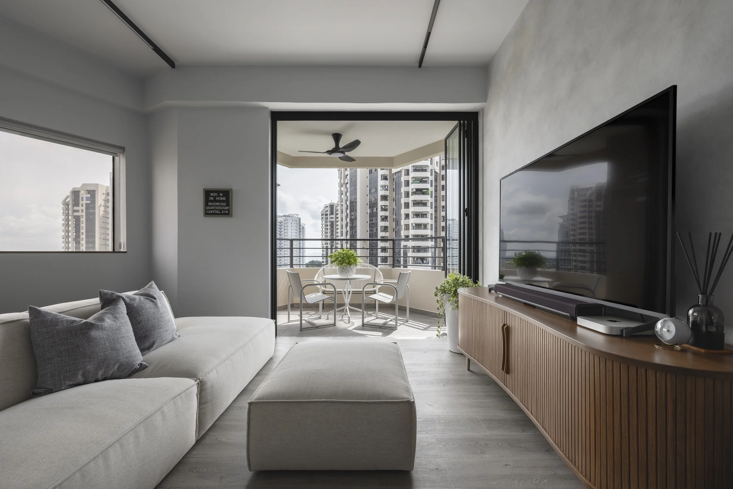 Modern living room with large white sofa, gray pillows, and a beige ottoman. A flat-screen TV is on a wooden media console. There is a small balcony with a round table, four chairs, and green plants. Tall buildings are visible outside.