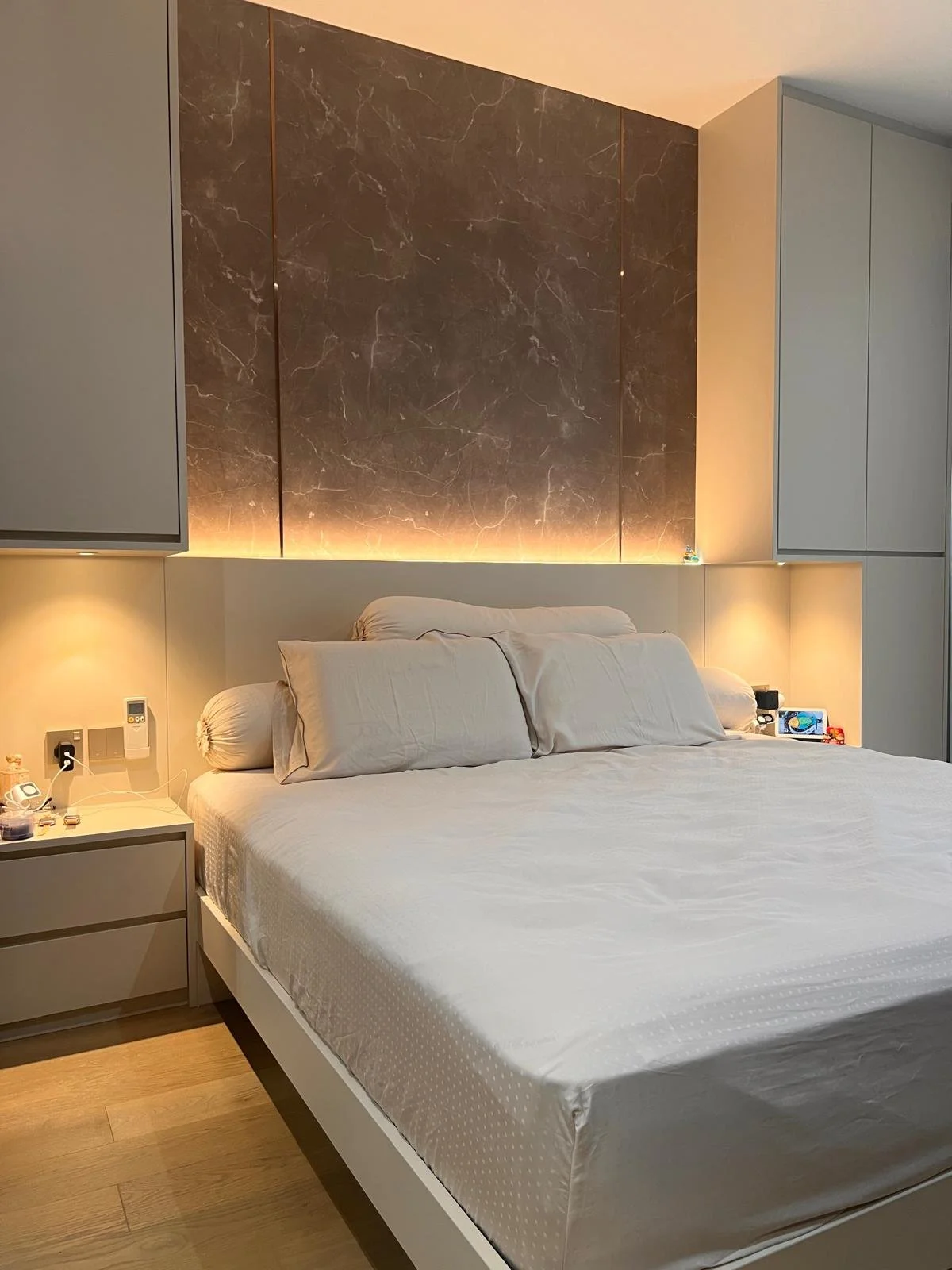 A neatly made bed with beige pillows and a white fitted sheet, in a modern bedroom with a marble accent wall and ambient lighting behind it. There are nightstands with small objects and a power outlet.