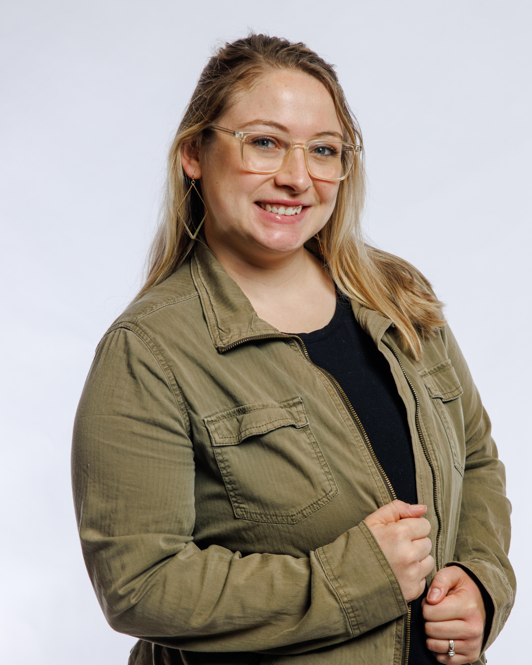 A young woman with blonde hair, glasses, and earrings, wearing an olive green jacket and black shirt, smiling and posing against a plain white background.