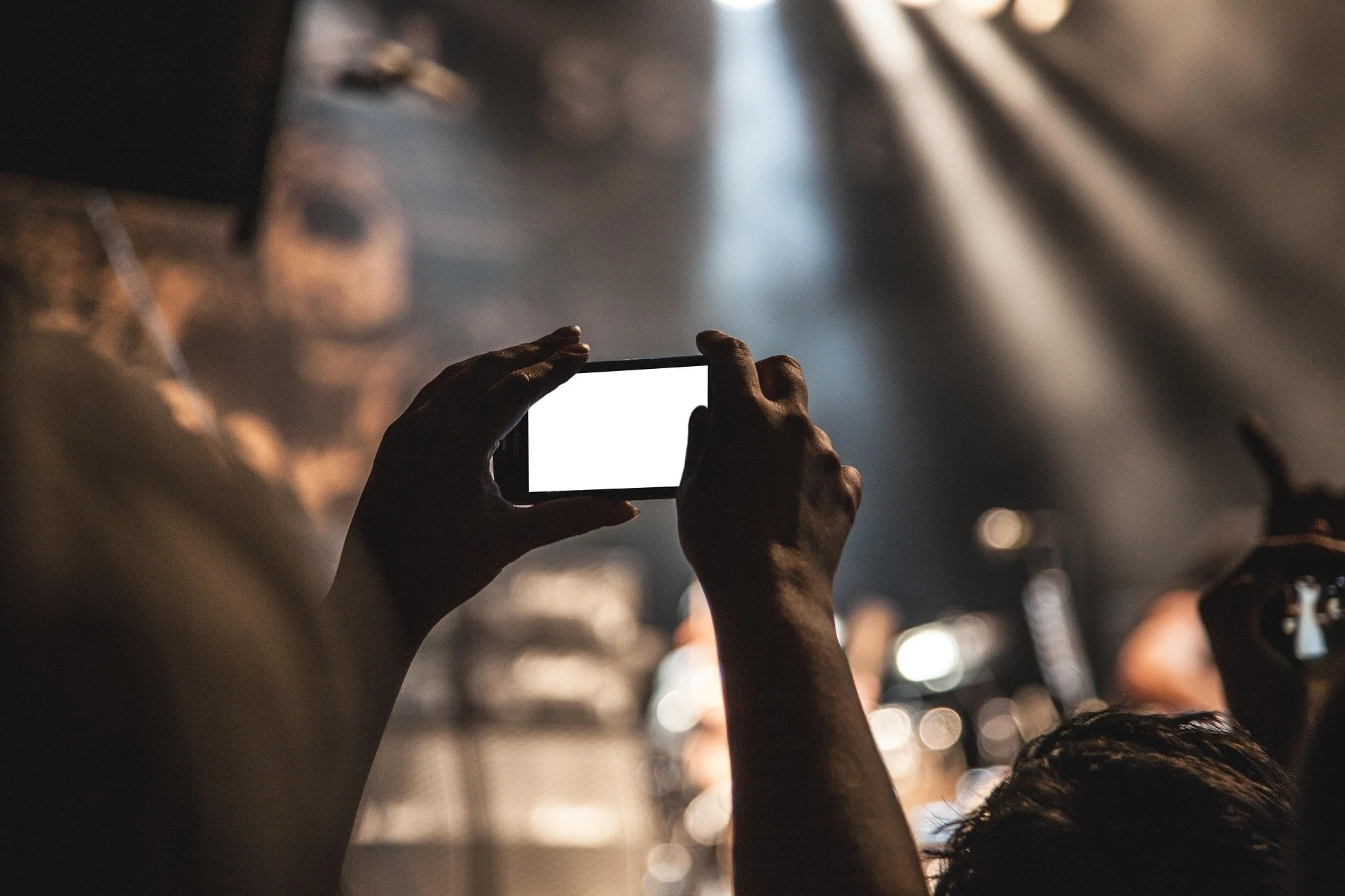 Person taking a photo with a smartphone at a concert or event, with stage lights and blurred crowd in the background.