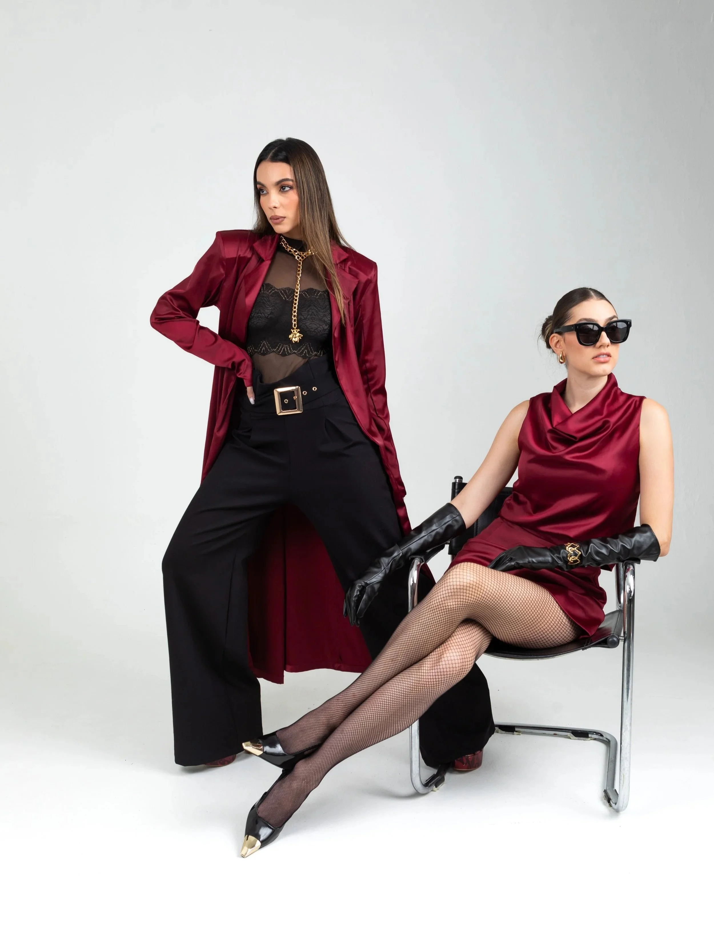 Two women in stylish maroon and black outfits, one sitting in a chair with black gloves and fishnet stockings, and the other standing with one hand on her hip, wearing sunglasses and jewelry, against a plain background.