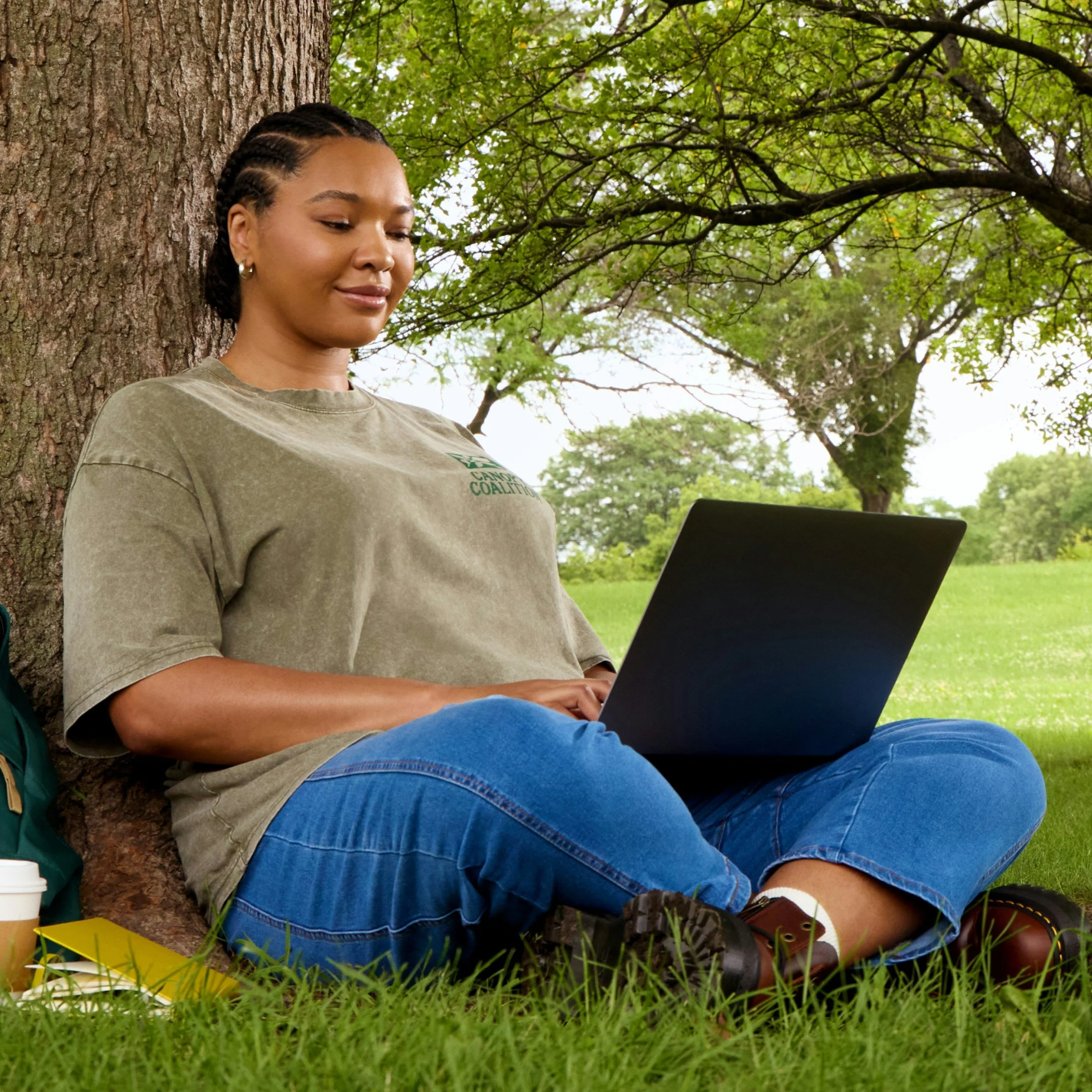 A woman sitting beneath a tree on grass, using a laptop, with a coffee cup and yellow notebook beside her.