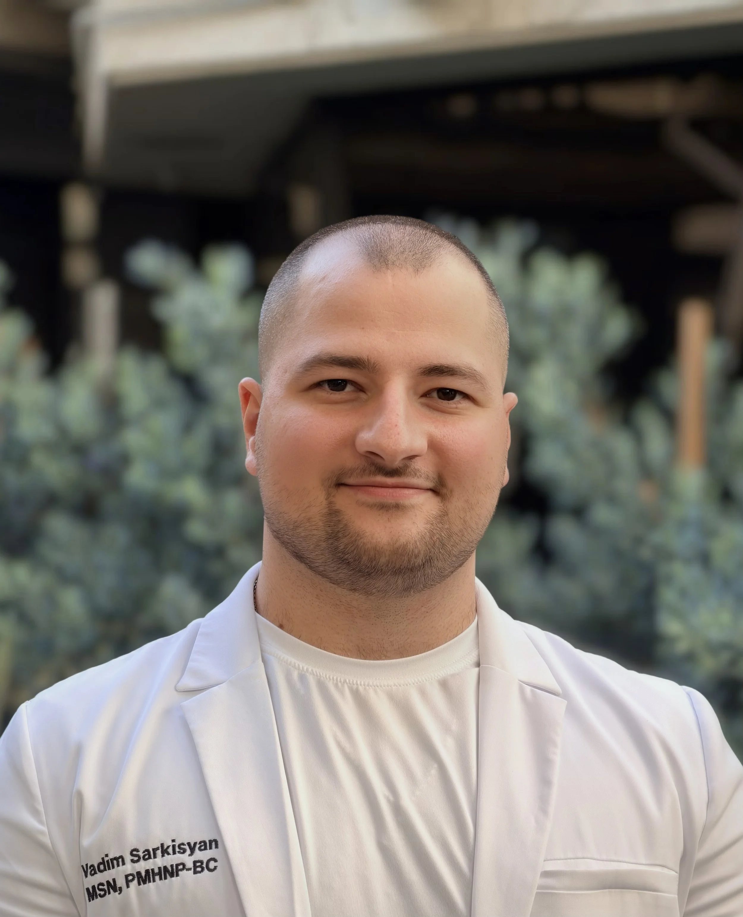 A young man in a white medical coat with a name tag that reads Vadim Sarkisyan, standing outdoors with a blurred natural background. PMHNP. Mental Health Services. Free Consultation.