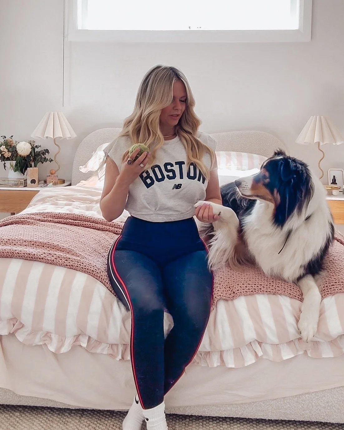 A woman sitting on a bed with a large dog, holding its paw. The woman is wearing a gray Boston T-shirt and blue athletic pants, holding a macaron. The bedroom has two lamps, flowers, and a window in the background.