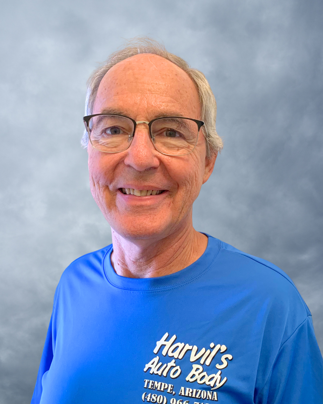 A smiling middle-aged man with glasses and light brown hair standing against a cloudy sky background, wearing a blue T-shirt with white text that reads 'Harvil's Auto Body, Tempe, Arizona, (480) 965-7...'