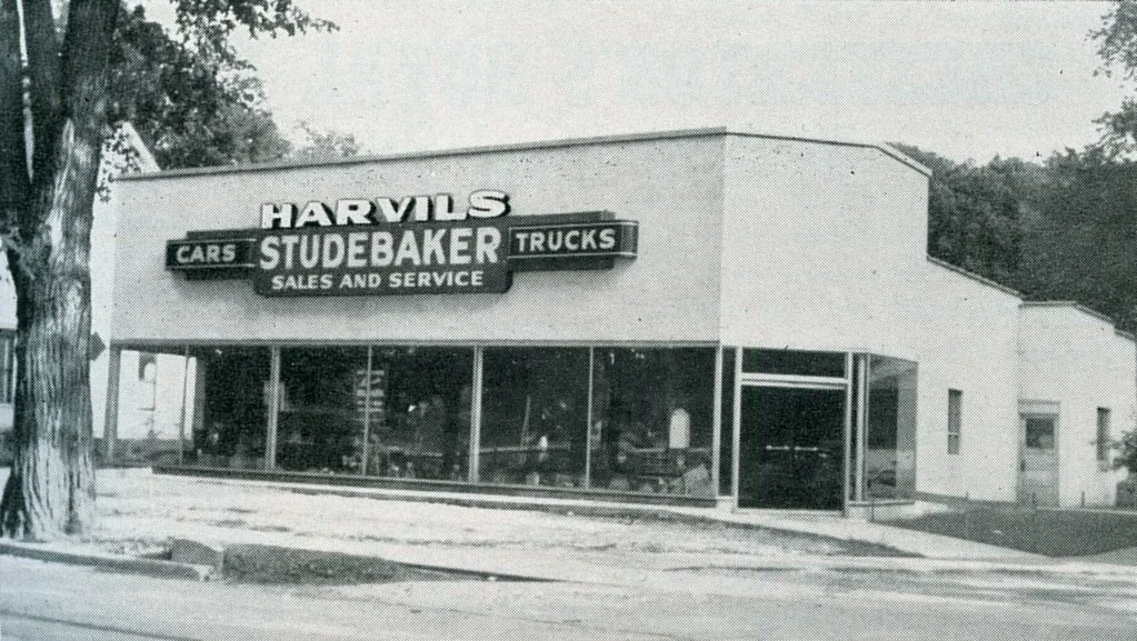A black and white photograph of a car dealership called Harvils Studebaker, featuring signage for cars, trucks, sales, and service. The building has large glass windows and is situated on a corner lot with a tree and sidewalk in front.