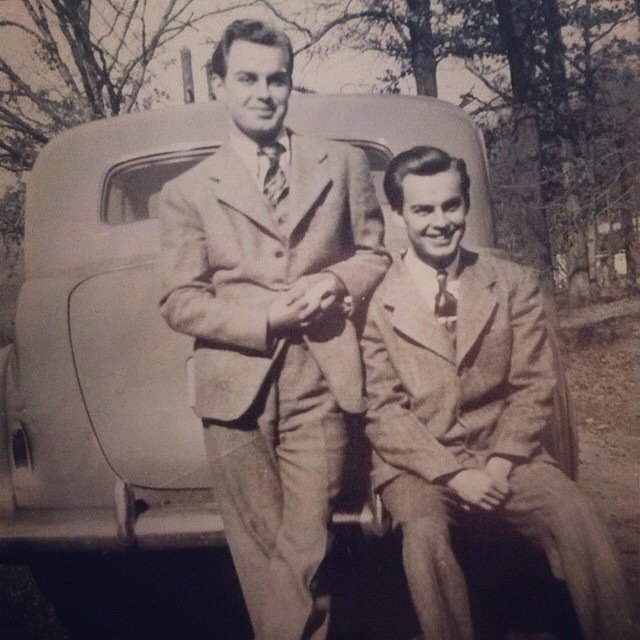 Two young men in vintage suits posing in front of a classic car outdoors, with trees and a house in the background.