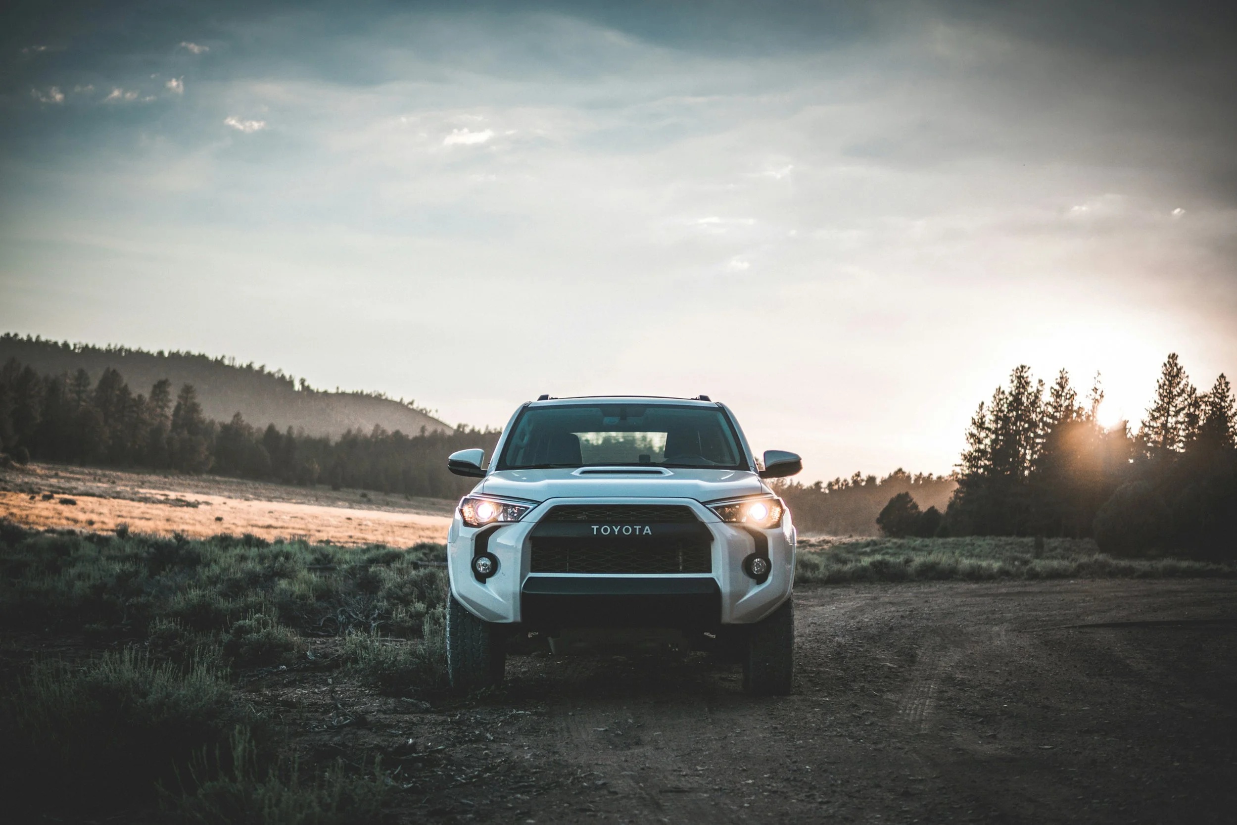 A white Toyota SUV parked on a dirt road in a rural landscape at sunset, with trees and hills in the background.