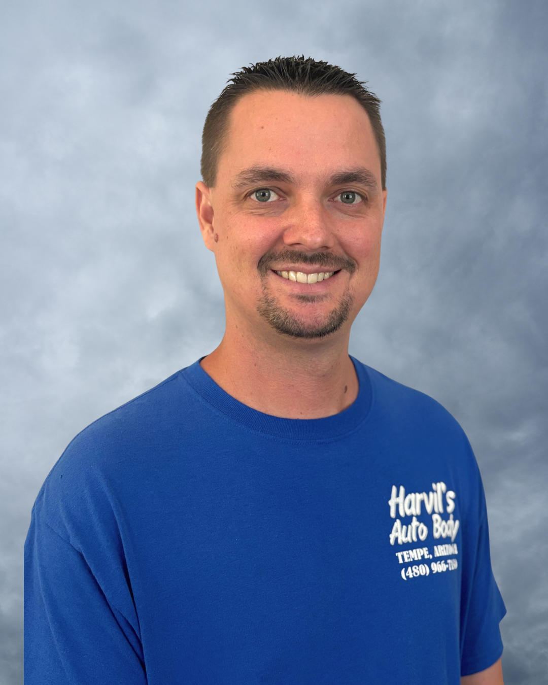 Smiling man with short dark hair and goatee, wearing a blue T-shirt with 'Harvil's Auto Body' and contact information, standing against a cloudy sky background.