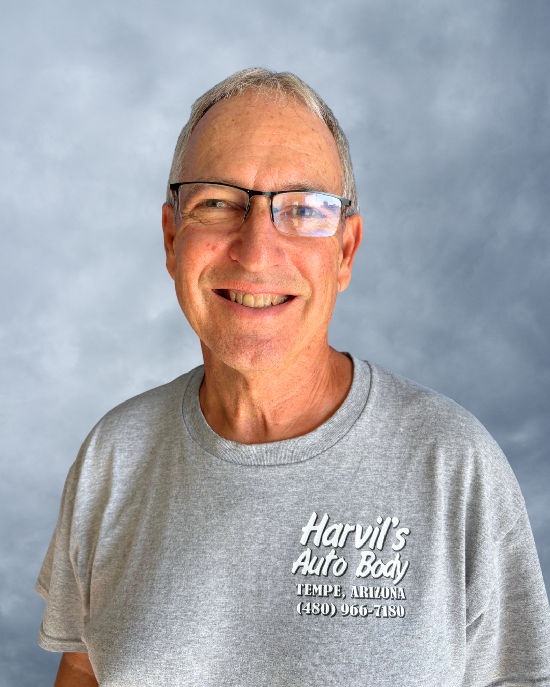 Smiling older man with glasses wearing a light gray 'Harvil's Auto Body' T-shirt with location and phone number, standing against a cloudy blue sky background.