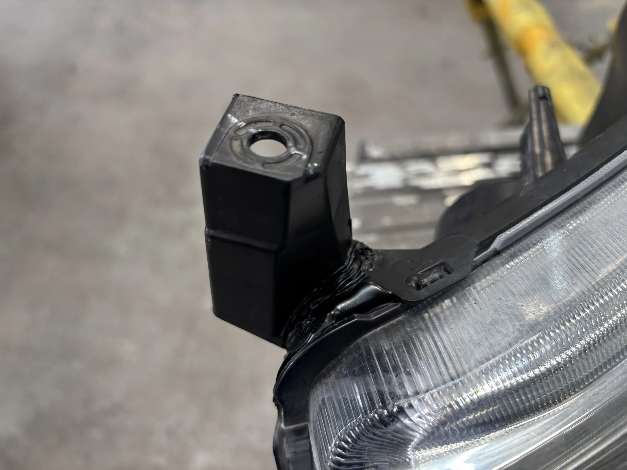 Close-up of a car headlight with a black mounting bracket or connector attached at the top, set against a garage or workshop background.