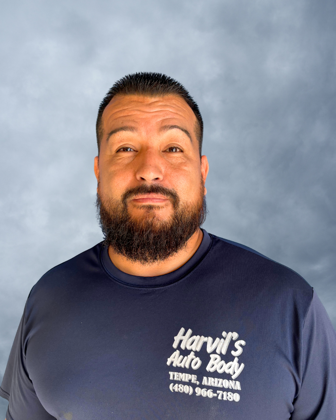 Portrait of a man with a beard wearing a navy blue t-shirt with the text "Havil's Auto Body, Tempe, Arizona, (480) 966-7180" against a gray cloudy background.