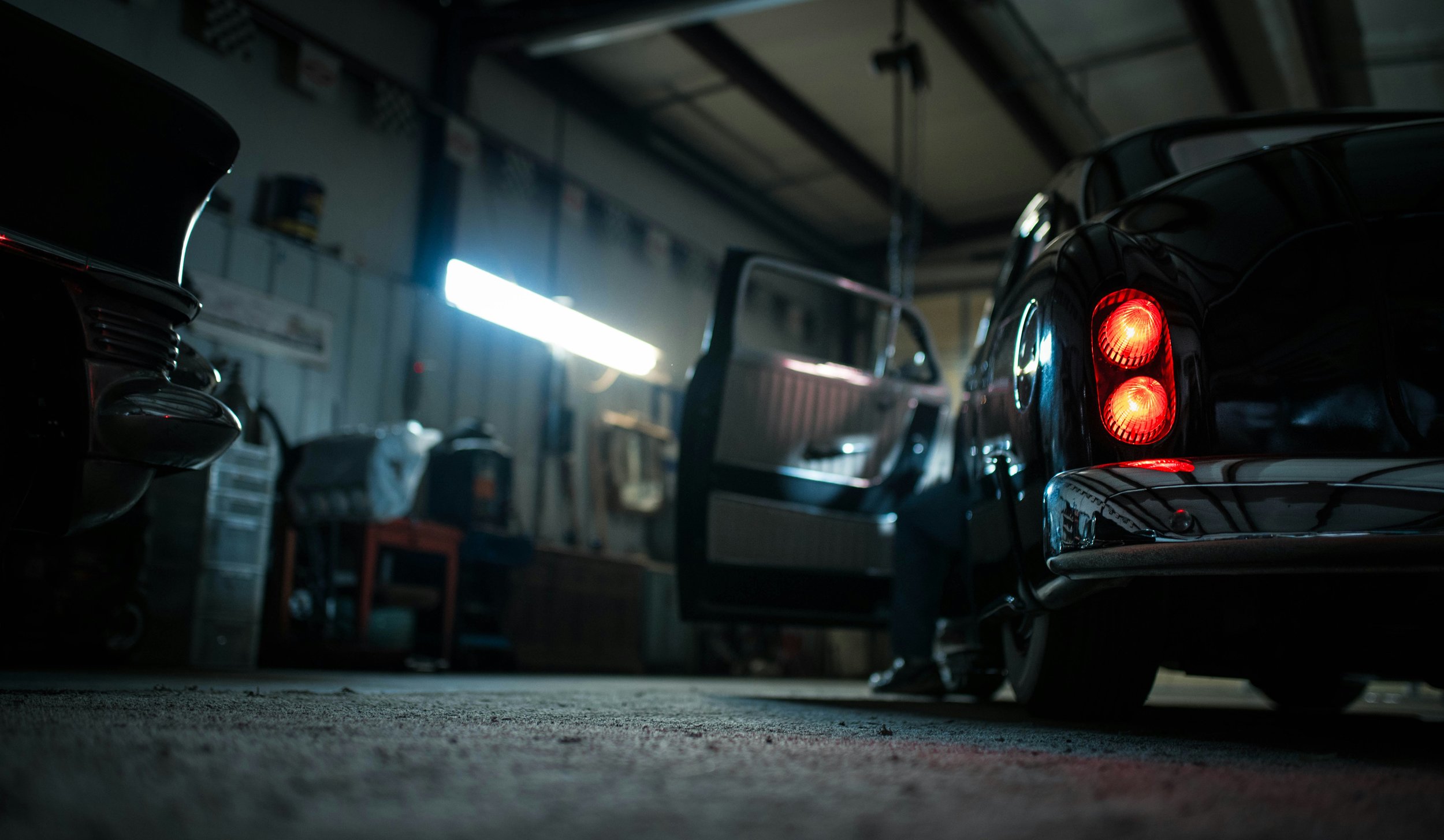 A garage with classic cars, including one with red taillights glowing, and open car doors. The scene is dimly lit with a bright fluorescent light on the wall.