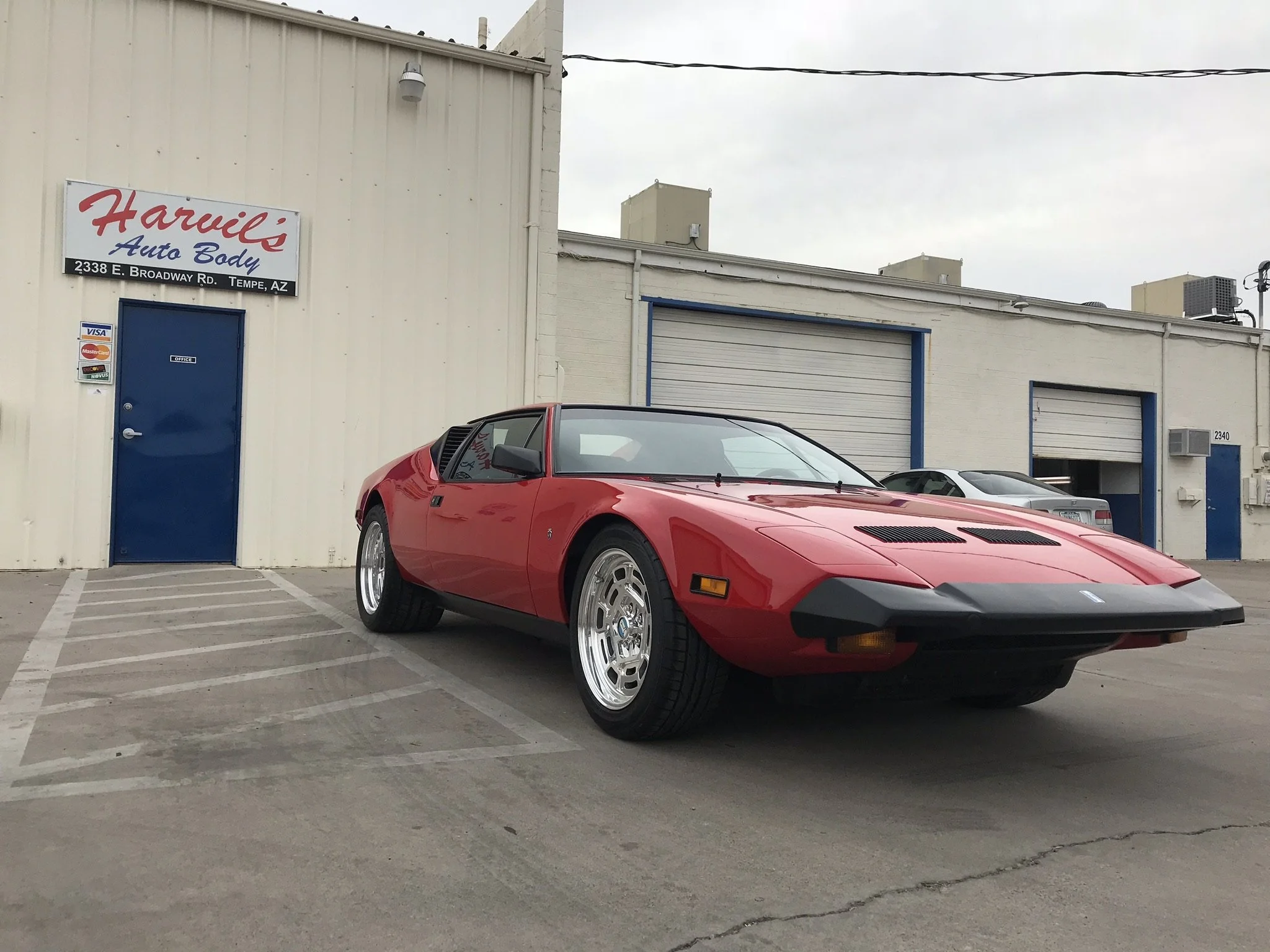 Red vintage sports car parked in front of an auto body shop with white building and blue doors.